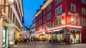 Una calle adoquinada flanqueada por edificios tradicionales europeos, incluyendo un hotel rojo con terraza y un restaurante.