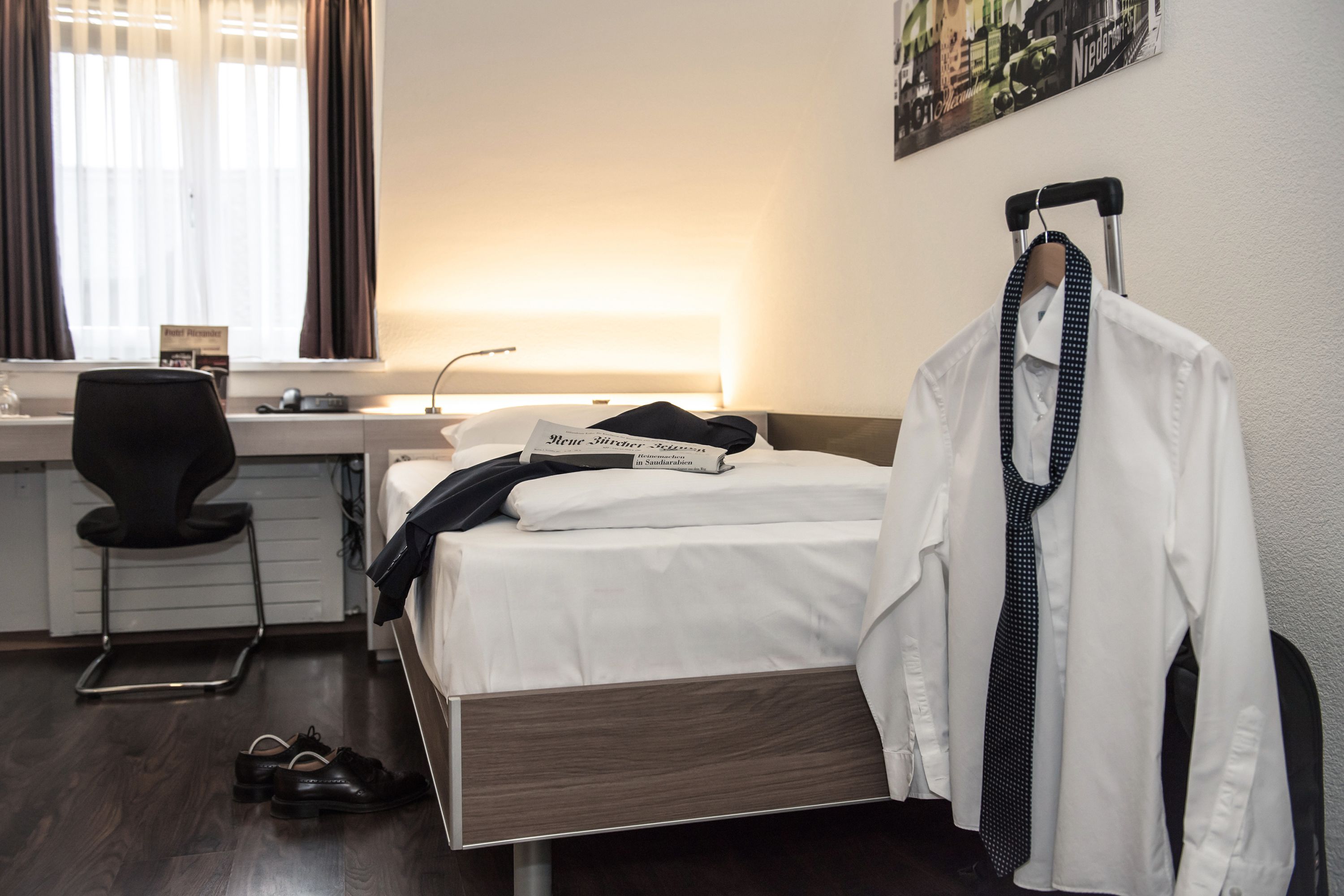 Quarto de hotel com cama, escrivaninha, cadeira, cabideiro e janela com cortinas.