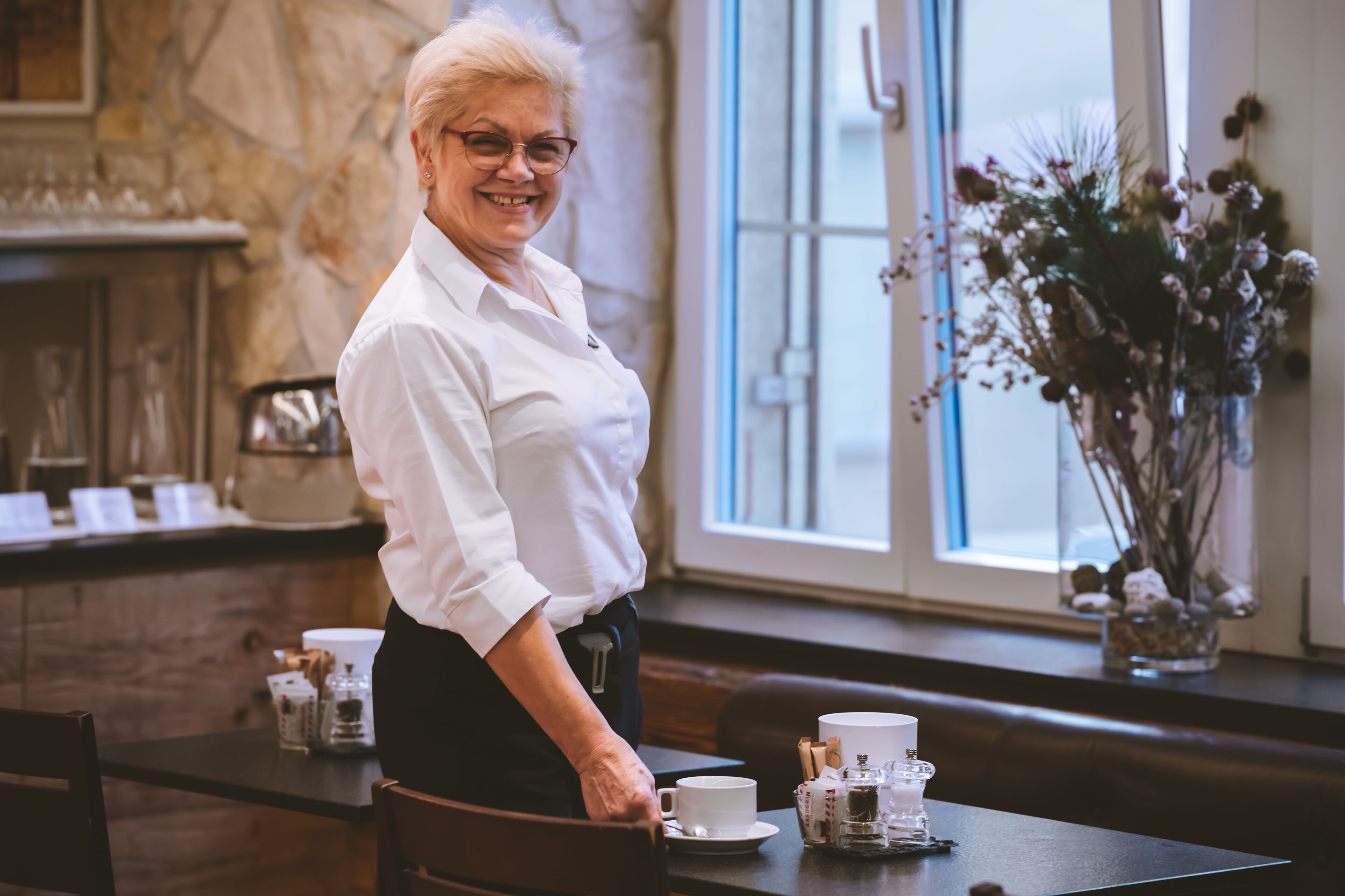 Una mujer de pie junto a una mesa con una preparación para café, un florero con flores secas y un muro de piedra al fondo.
