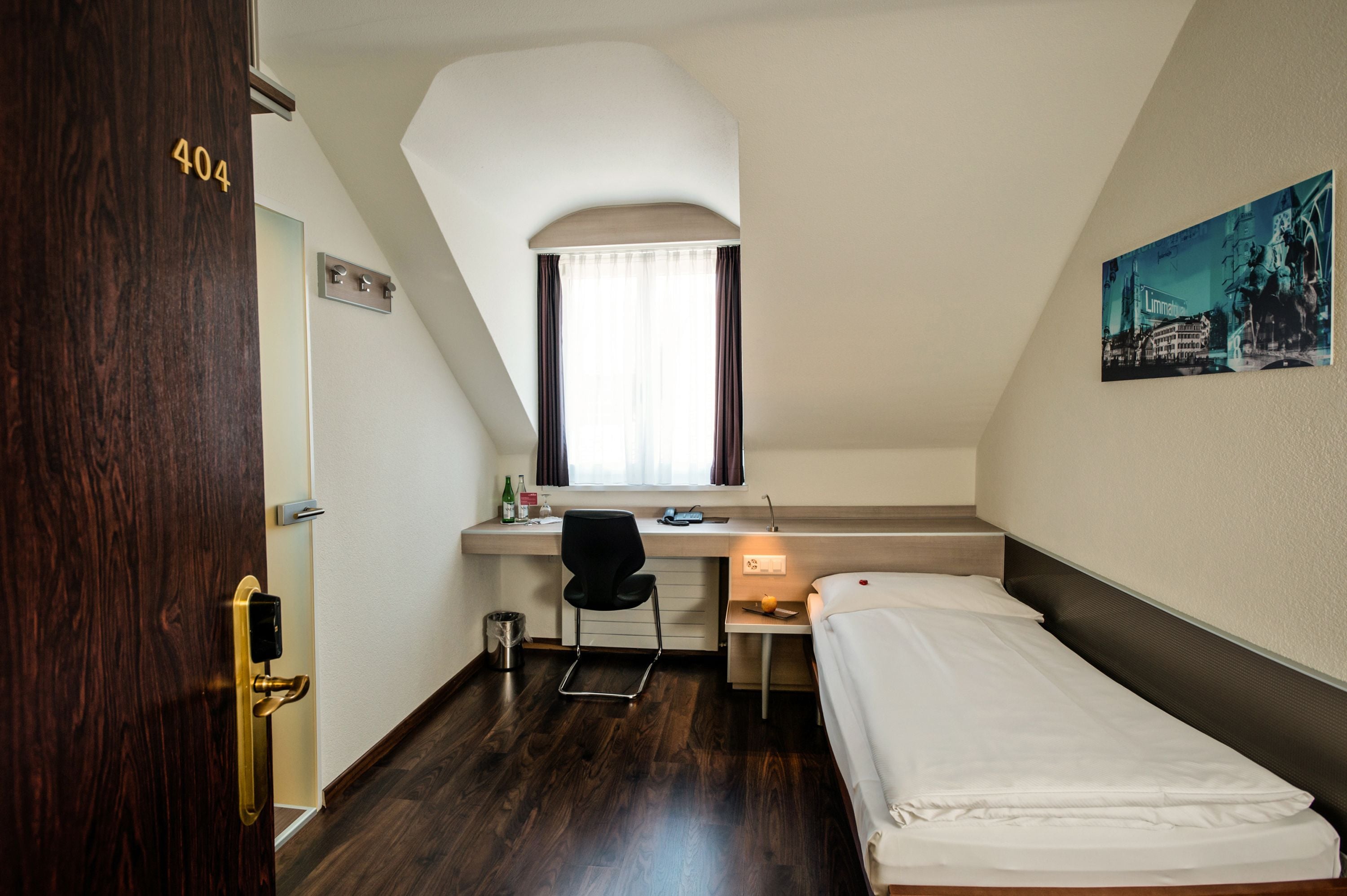 A small, modern hotel room with a single bed, a desk with a chair, a window with curtains, and a door with a number.