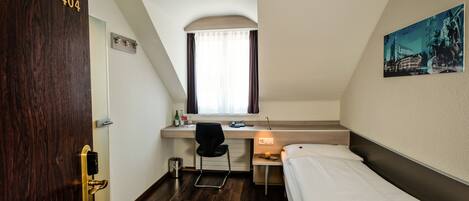 A small, modern hotel room with a single bed, a desk with a chair, a window with curtains, and a door with a number.