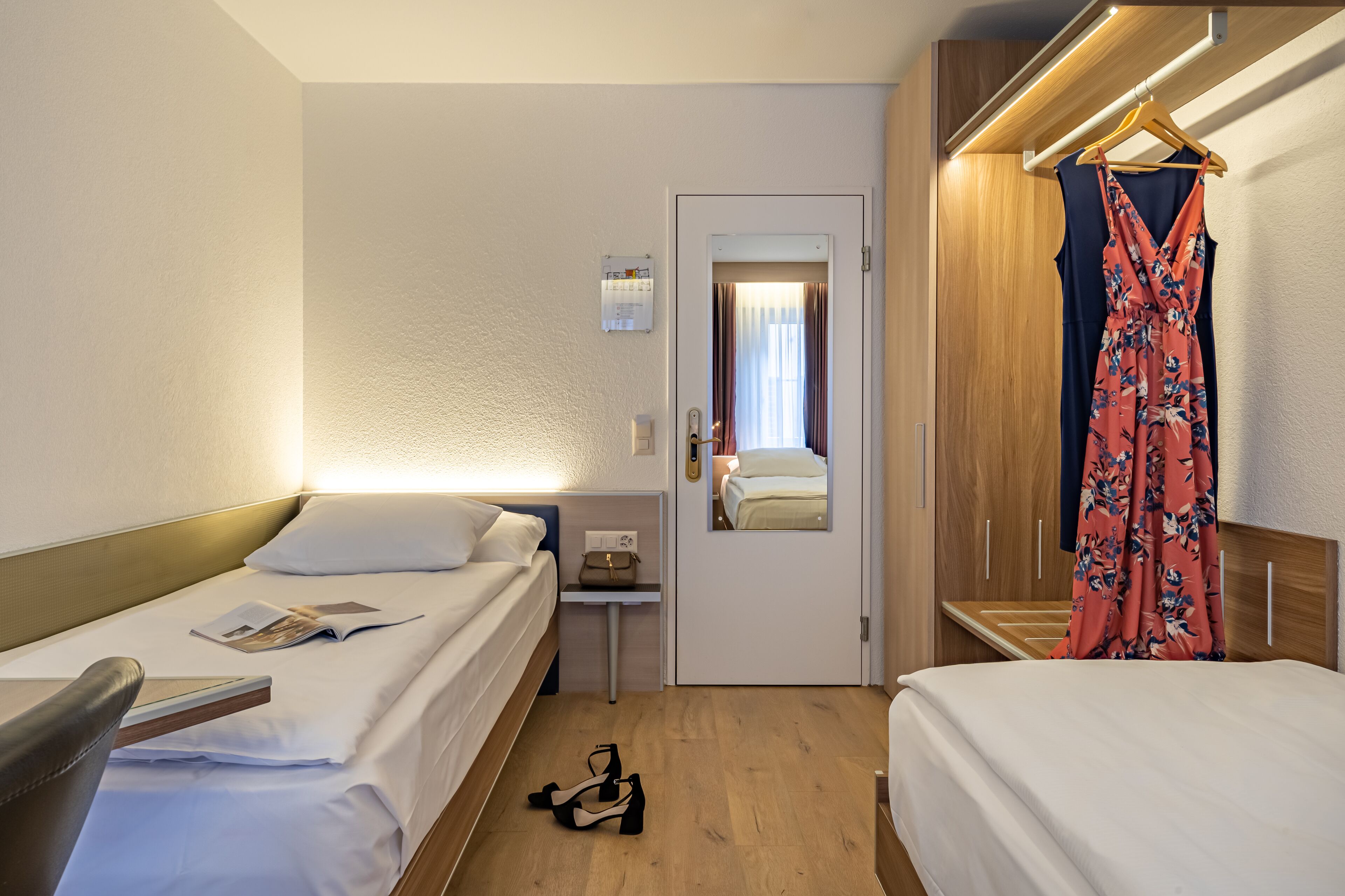 Habitación de hotel con dos camas, una mesita de noche, un espejo, un vestido colgado y un par de zapatos en el suelo.