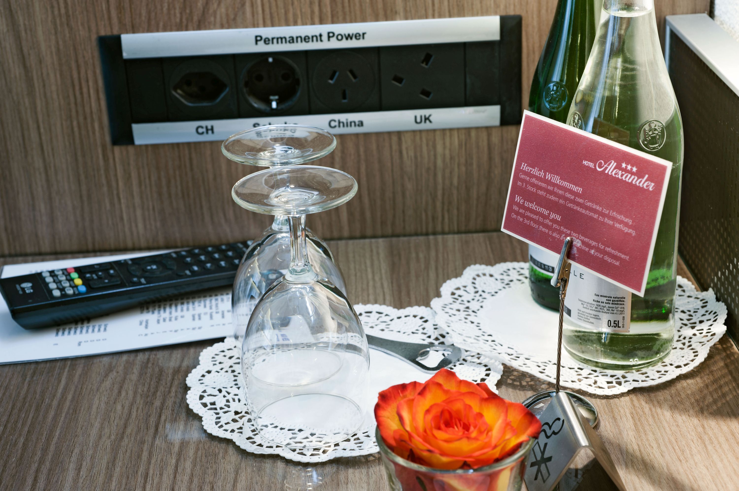 Une chambre d’hôtel avec une télécommande, un verre à vin, une rose et une bouteille de vin posés sur une table.