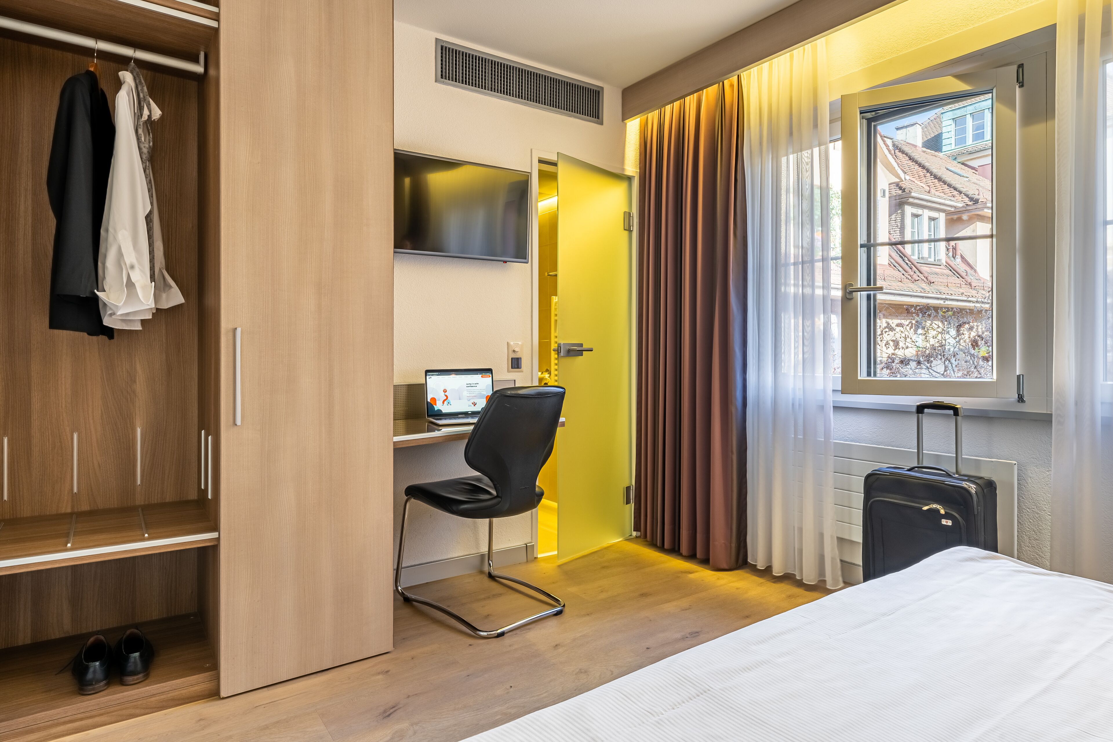 Ein Hotelzimmer mit einem Bett, einem Schreibtisch mit Computer, einem Stuhl, einem Kleiderschrank und einem Koffer.