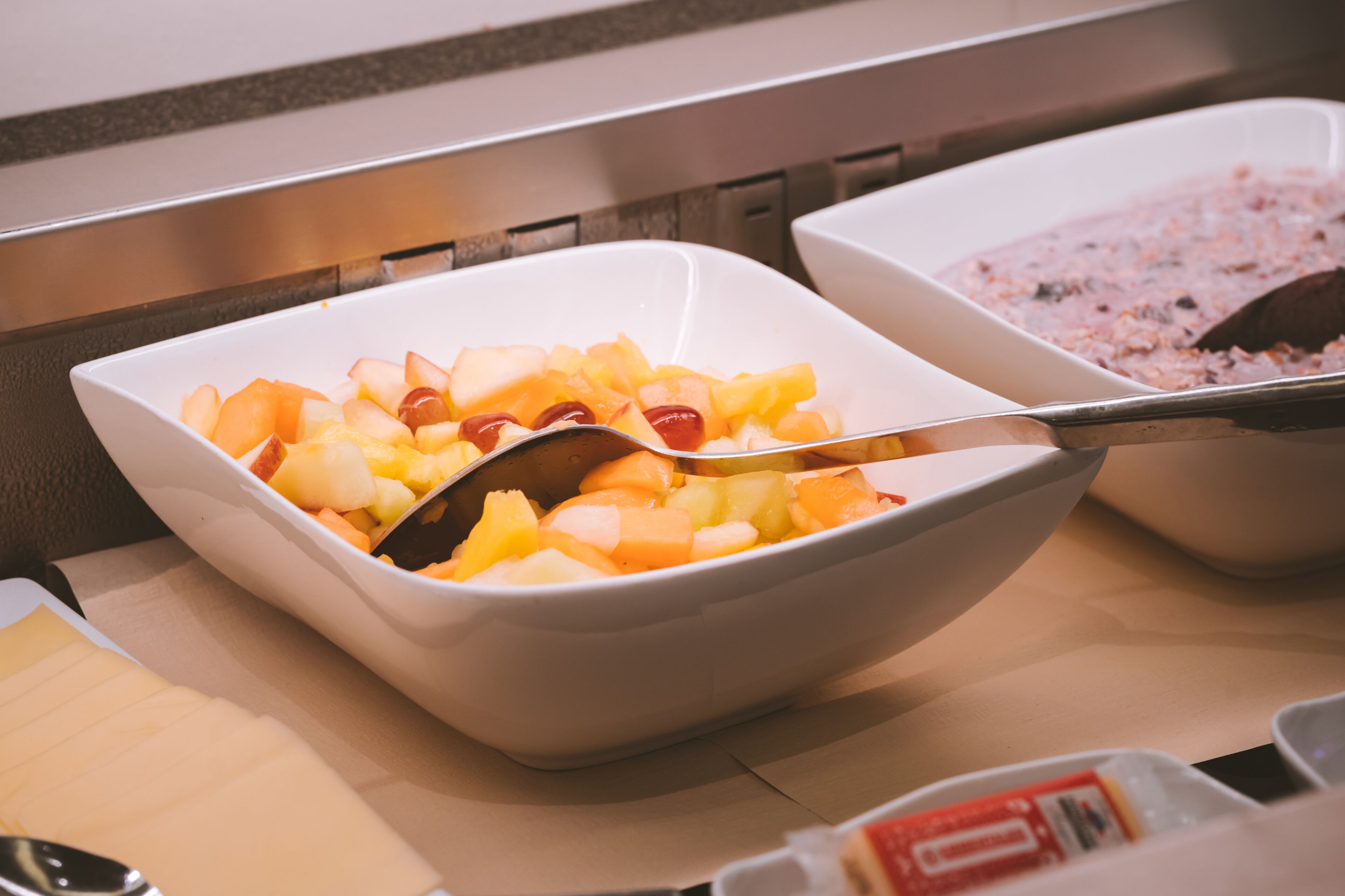 Un tazón de ensalada de frutas con una cuchara, sobre una mesa con servilleta y condimentos.