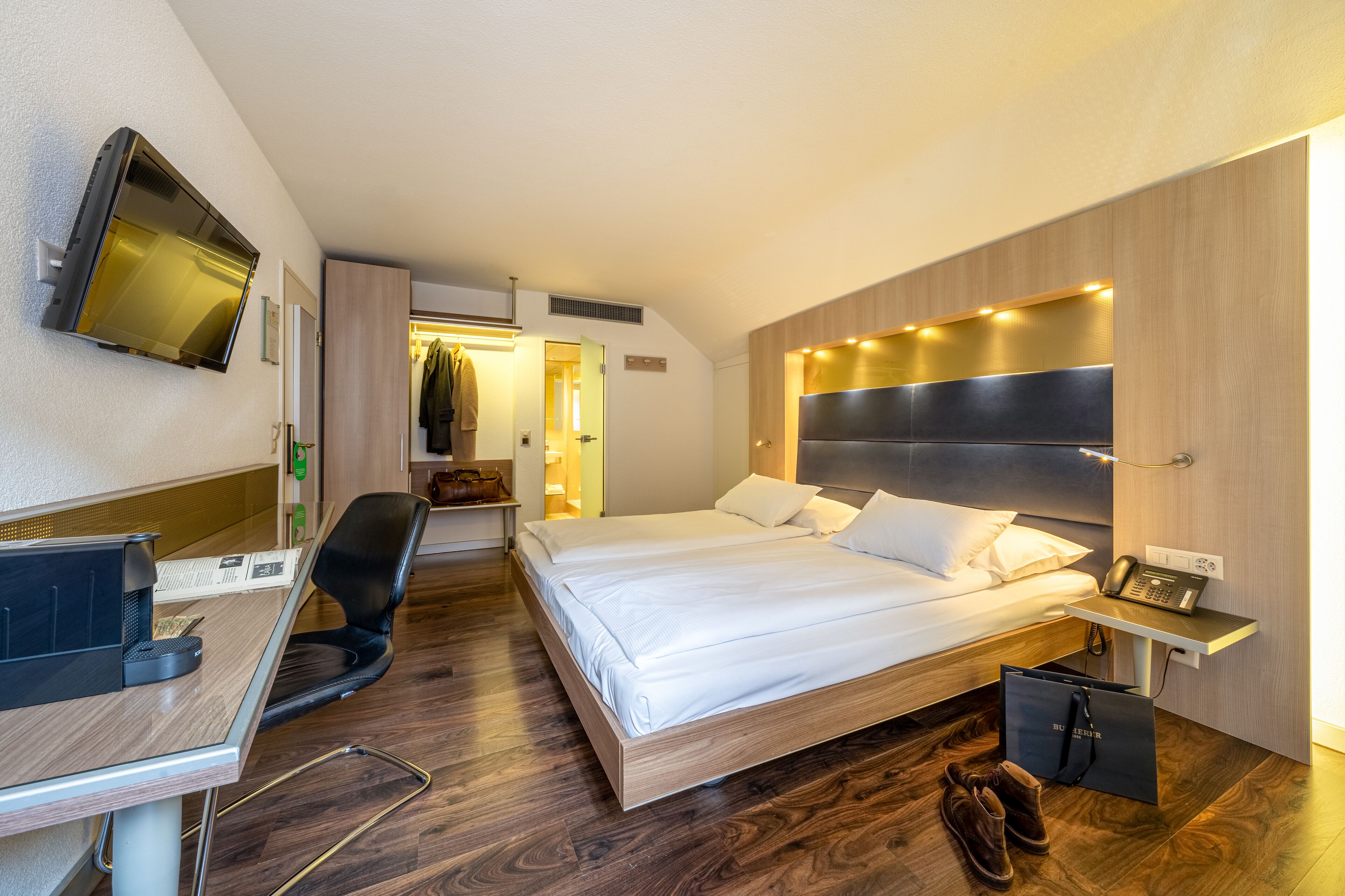 Una habitación de hotel moderna con una cama, escritorio, silla y televisor instalado en la pared.