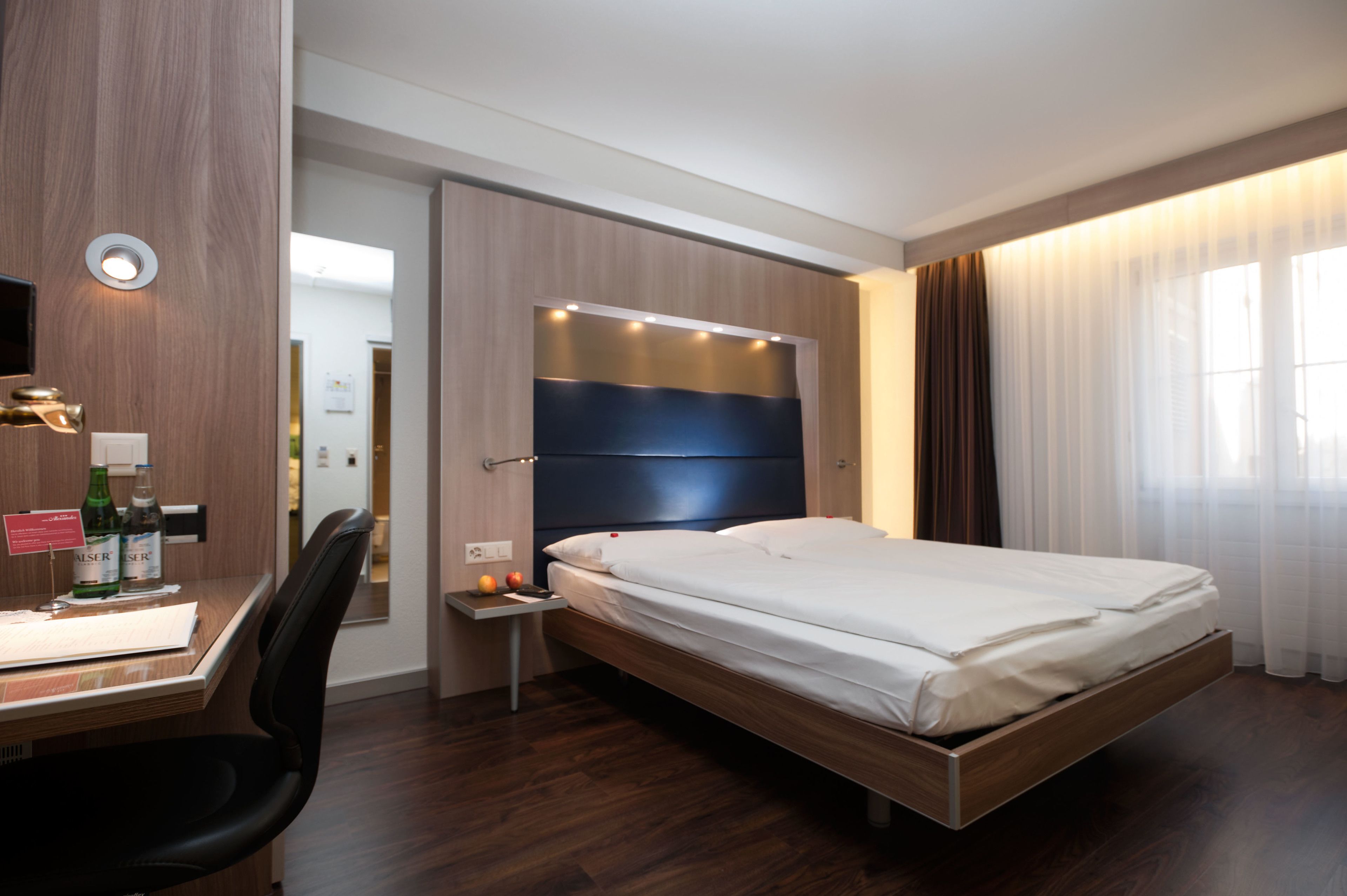 A modern hotel room with a wooden bed frame, a desk with a chair, a nightstand with a lamp, and a large window with curtains.