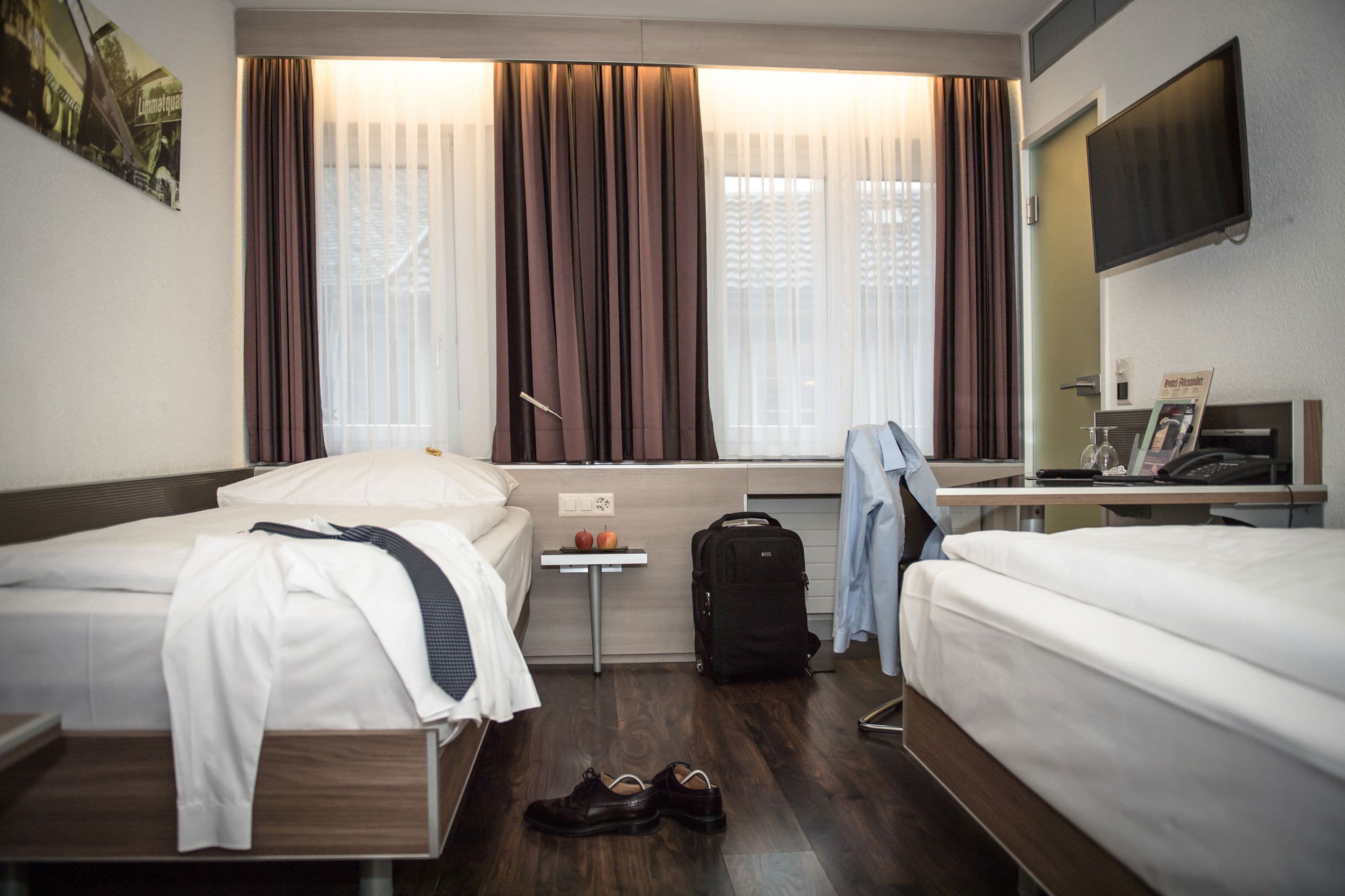 A hotel room with two beds, a small table with a fruit bowl, a suitcase, and a TV on the wall.