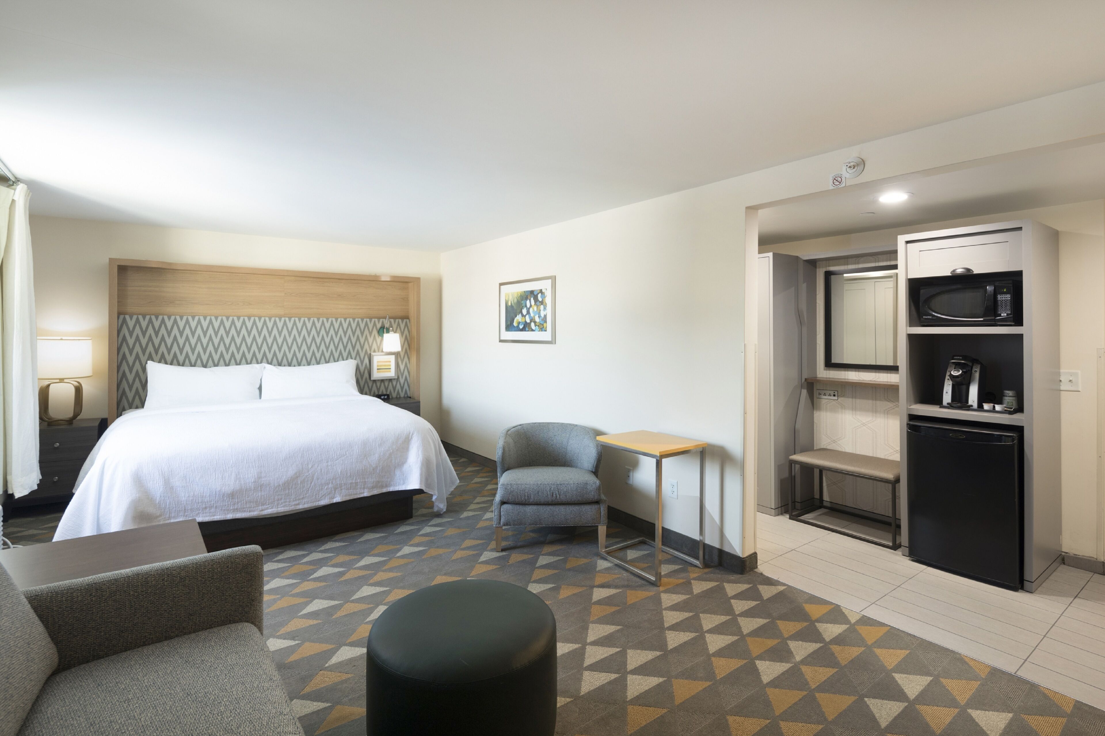 A hotel room with a bed, a small desk, a chair, a sofa, a bench, and a kitchenette.