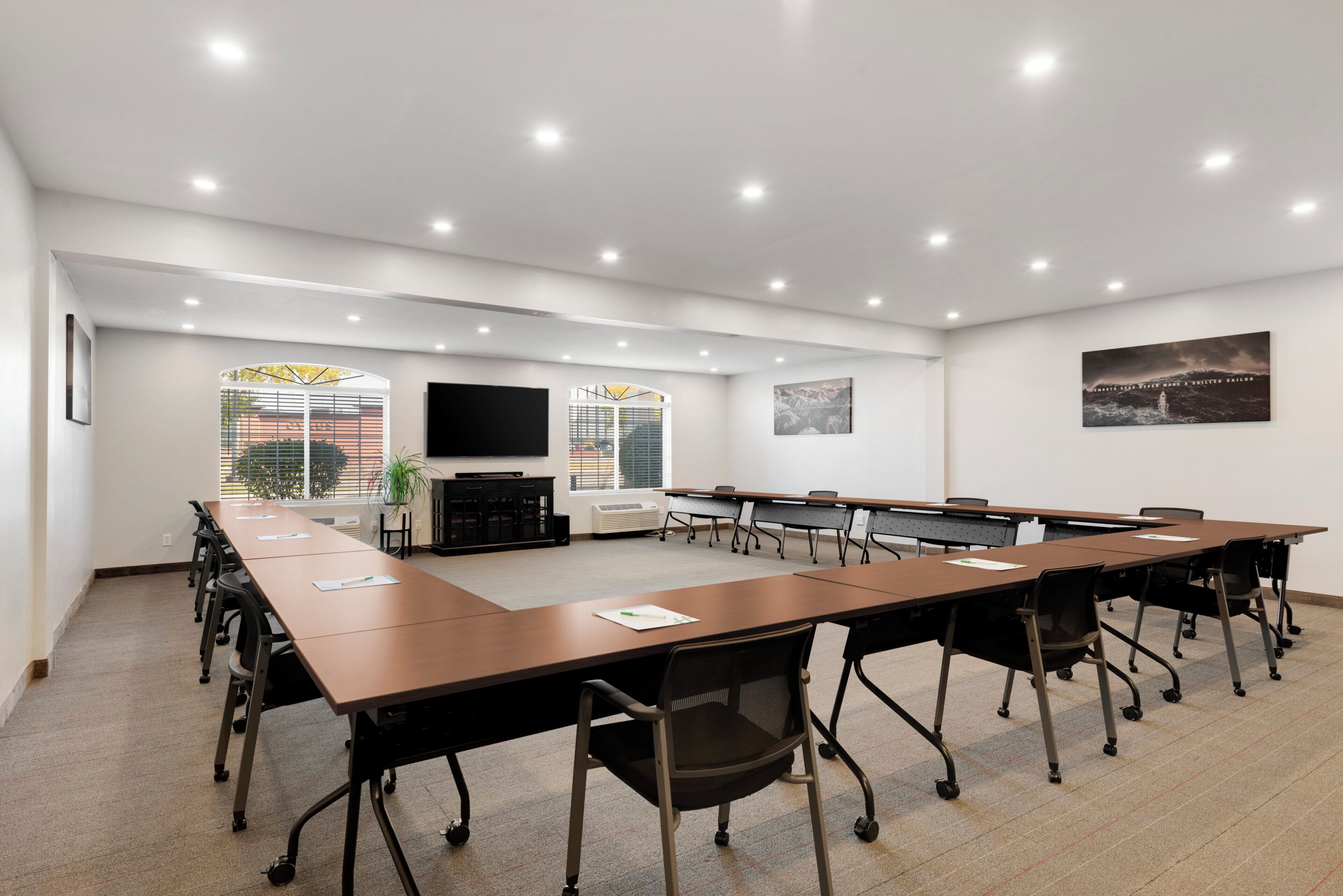 Een congreszaal met een lange tafel, stoelen, een flatscreen televisie en een groot raam met jaloezieën.