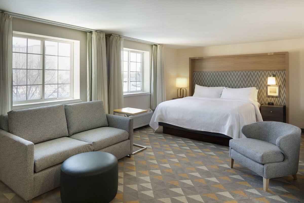 A hotel room with a bed, a sofa, a chair, a small table, and a lamp.