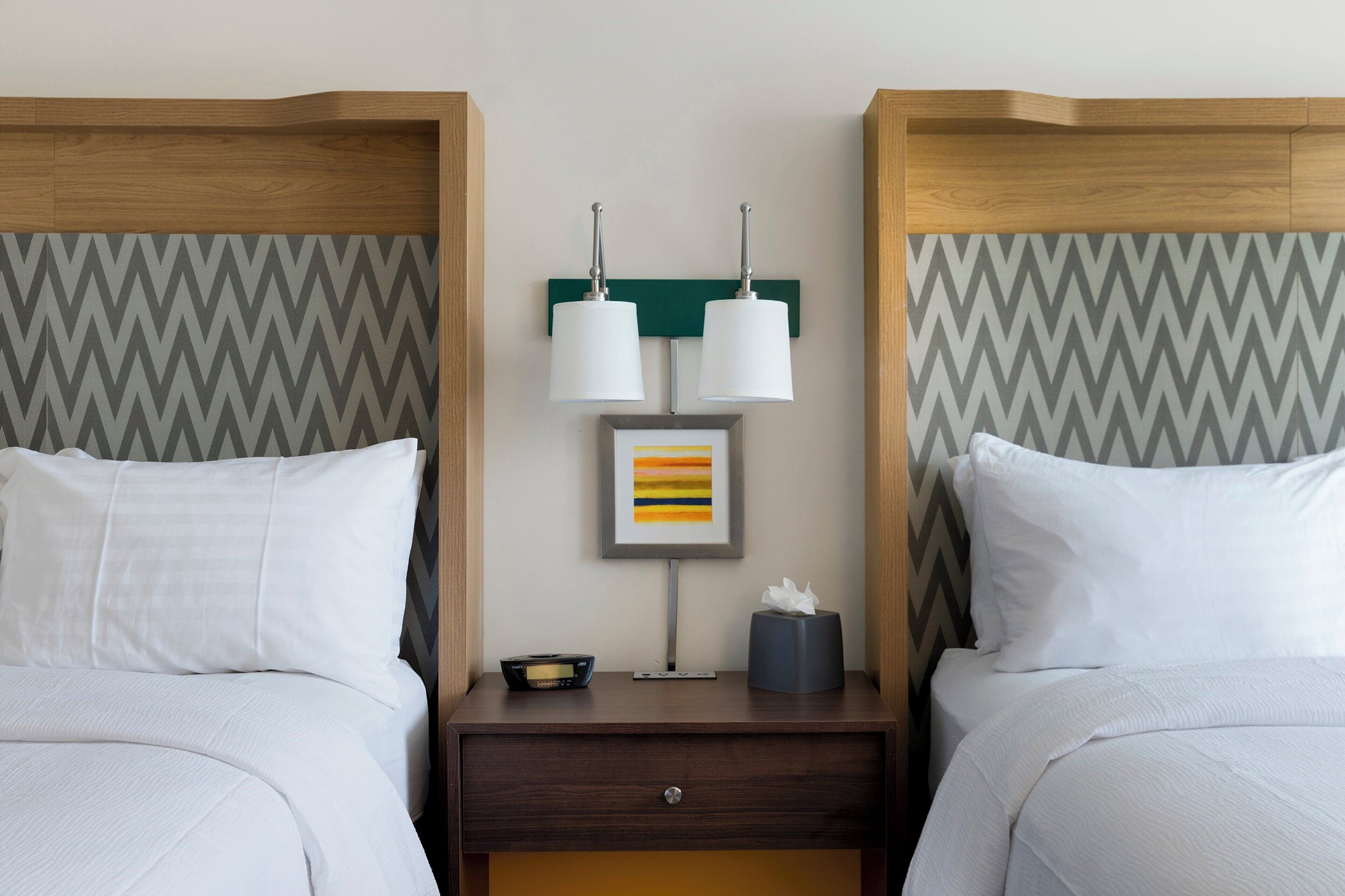 Habitación de hotel con dos camas, una mesita de noche con lámpara, un cuadro enmarco y una caja de pañuelos.