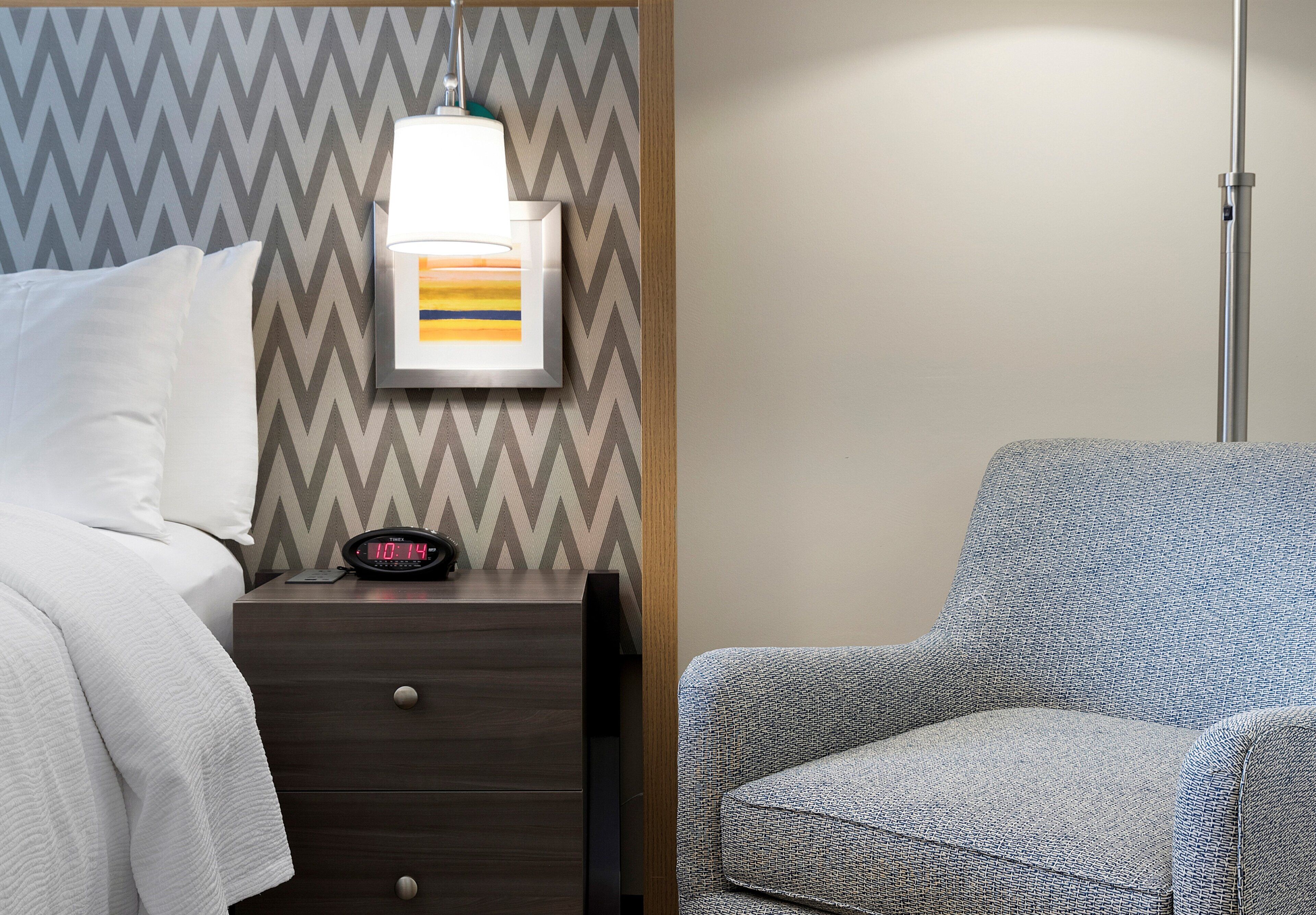 A hotel room with a bed, a nightstand, a digital clock, a lamp, and a patterned armchair.