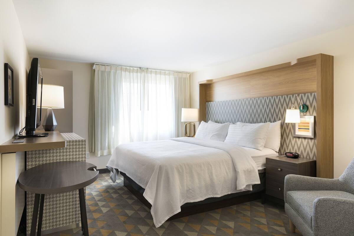 A hotel room with a bed, a desk, a chair, a nightstand, and a lamp.
