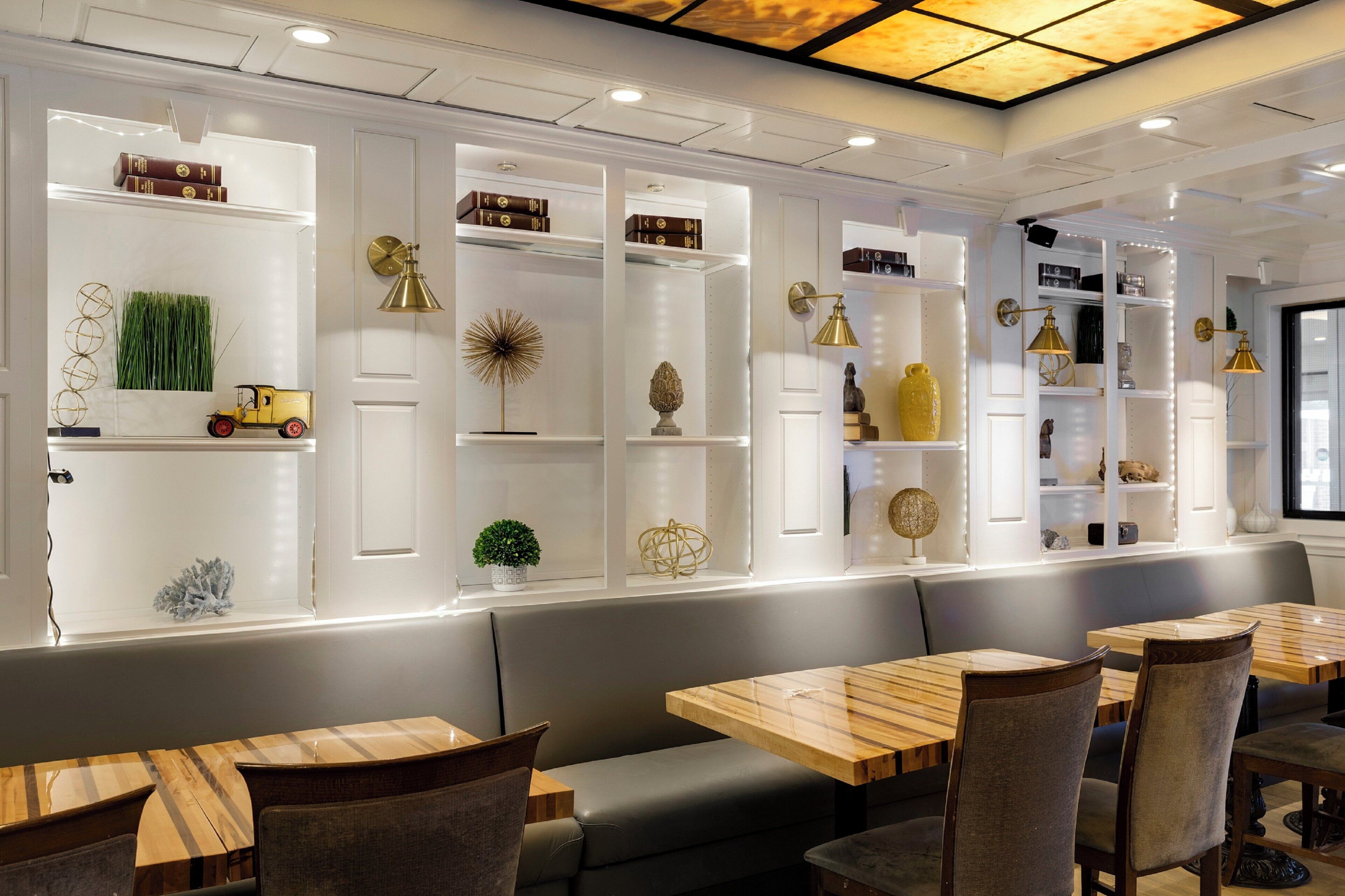 Ein Restaurantinnenraum mit Holztischen und -stühlen, weißen Regalen mit Dekoration und einer Decke mit eingelassener Beleuchtung.