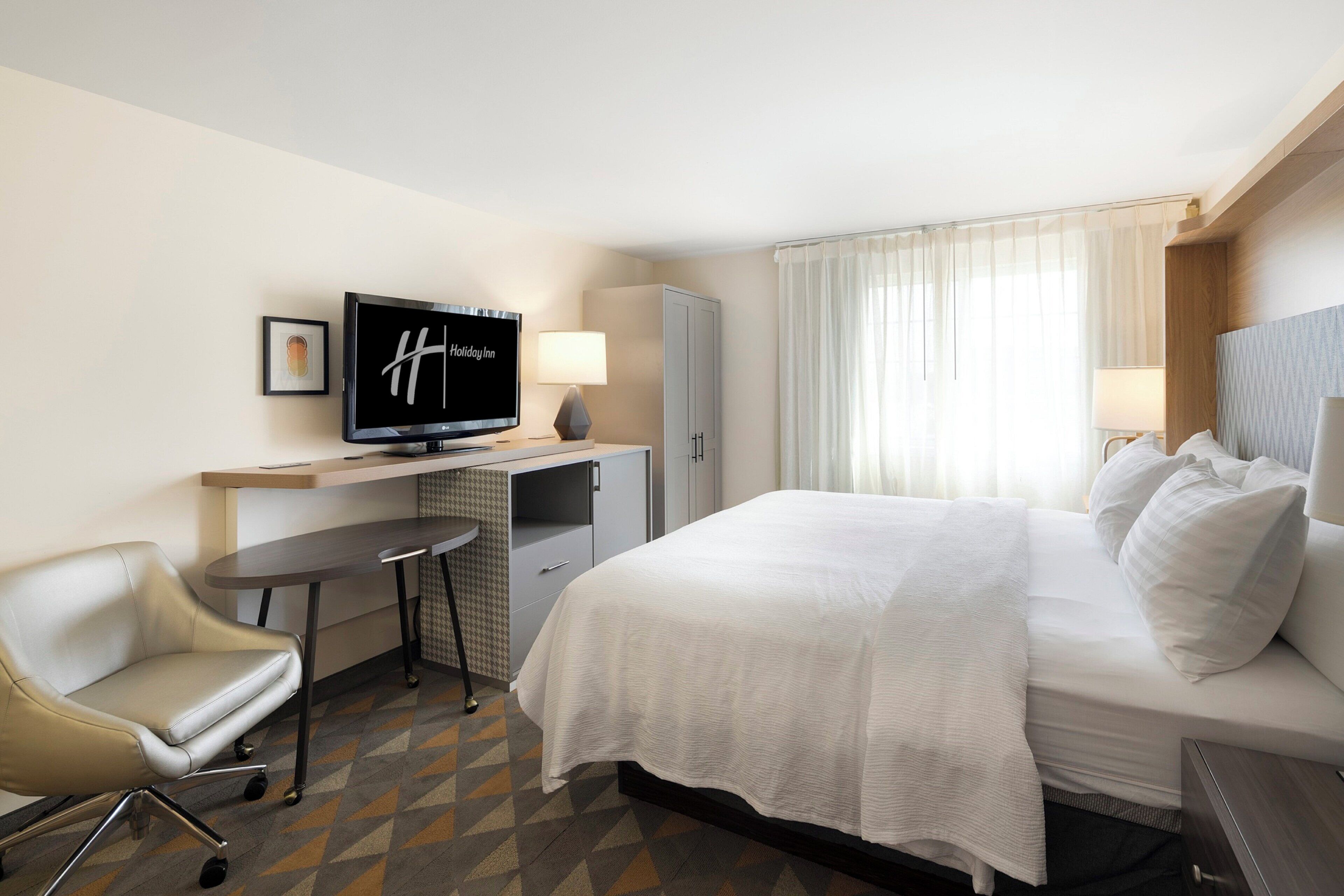 Une chambre d’hôtel avec un lit, un bureau avec une télévision, une chaise et une fenêtre avec des rideaux.