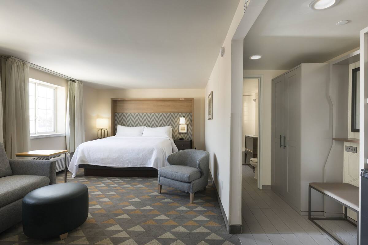 A hotel room with a bed, a chair, a desk, a wardrobe, and a bathroom visible through an open door.