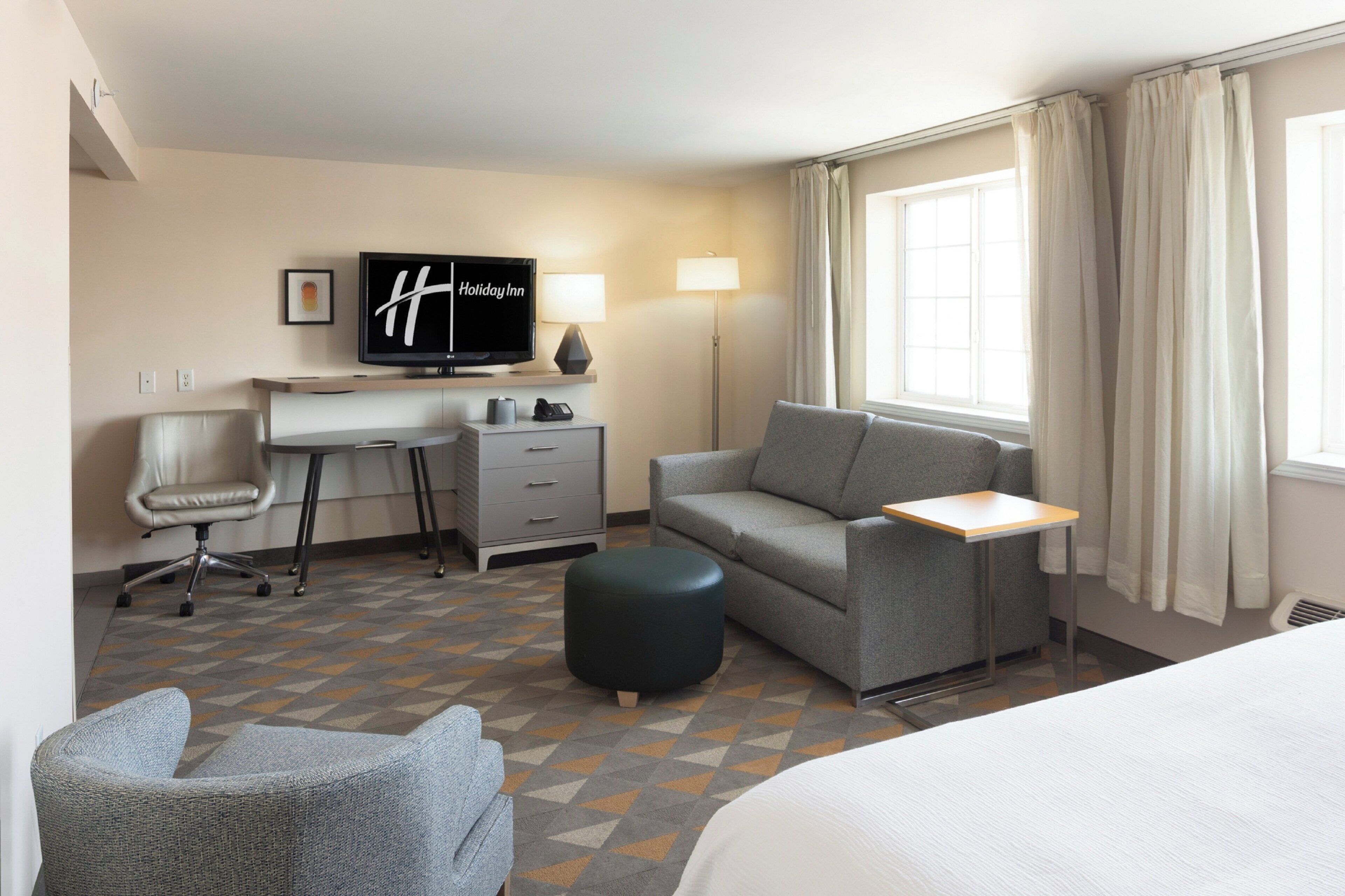 Habitació d'hotel amb un llit, un escriptori, una cadira, un sofà, una televisió, una làmpada i una finestra amb cortines.
