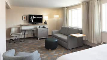 Habitació d'hotel amb un llit, un escriptori, una cadira, un sofà, una televisió, una làmpada i una finestra amb cortines.