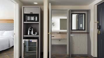 Habitación de hotel con cama, minibar, cafetera, refrigerador, baño con lavamanos y espejo, y una puerta.
