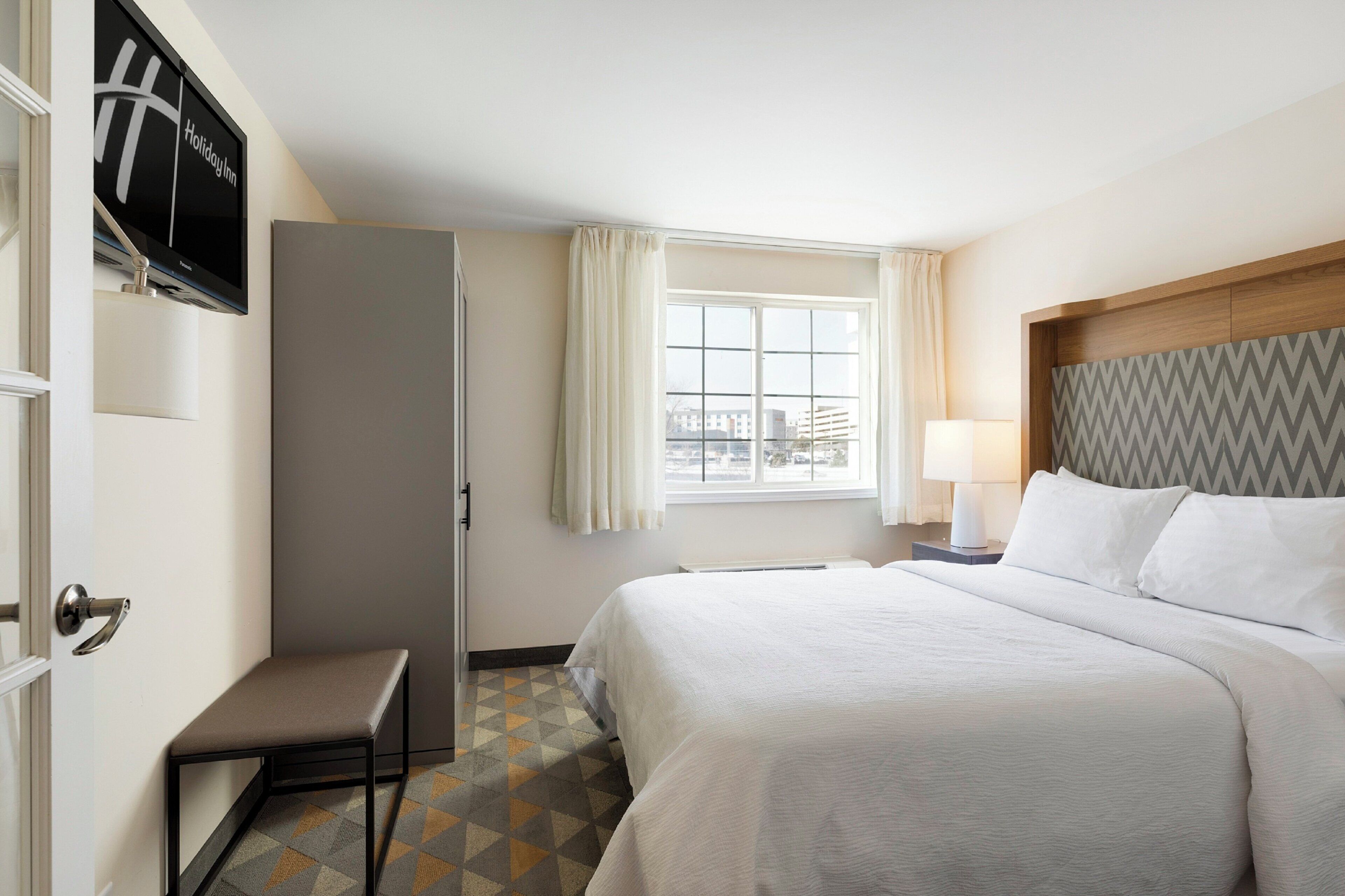Una habitación de hotel con una cama, un banco, una ventana con cortinas, un televisor en la pared y un piso con diseño.