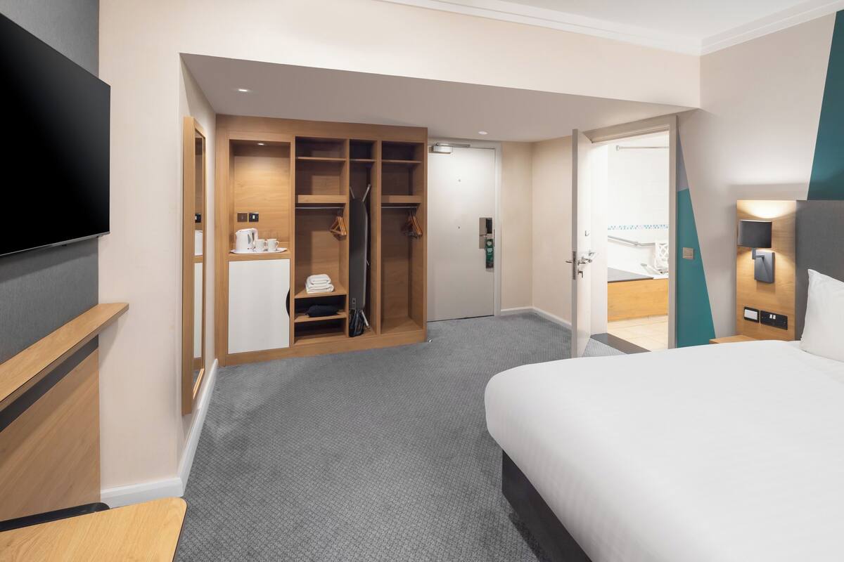 Ein Hotelzimmer mit einem großen Bett, einem an der Wand befestigten Fernseher, einem Schrank mit Spiegel und einer kleinen Küchenzeile.