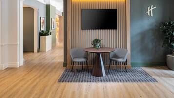 Un lobby moderno con una mesa redonda de madera, dos sillas grises, una planta en maceta y un televisor de pantalla plana montado en una pared de paneles de madera.