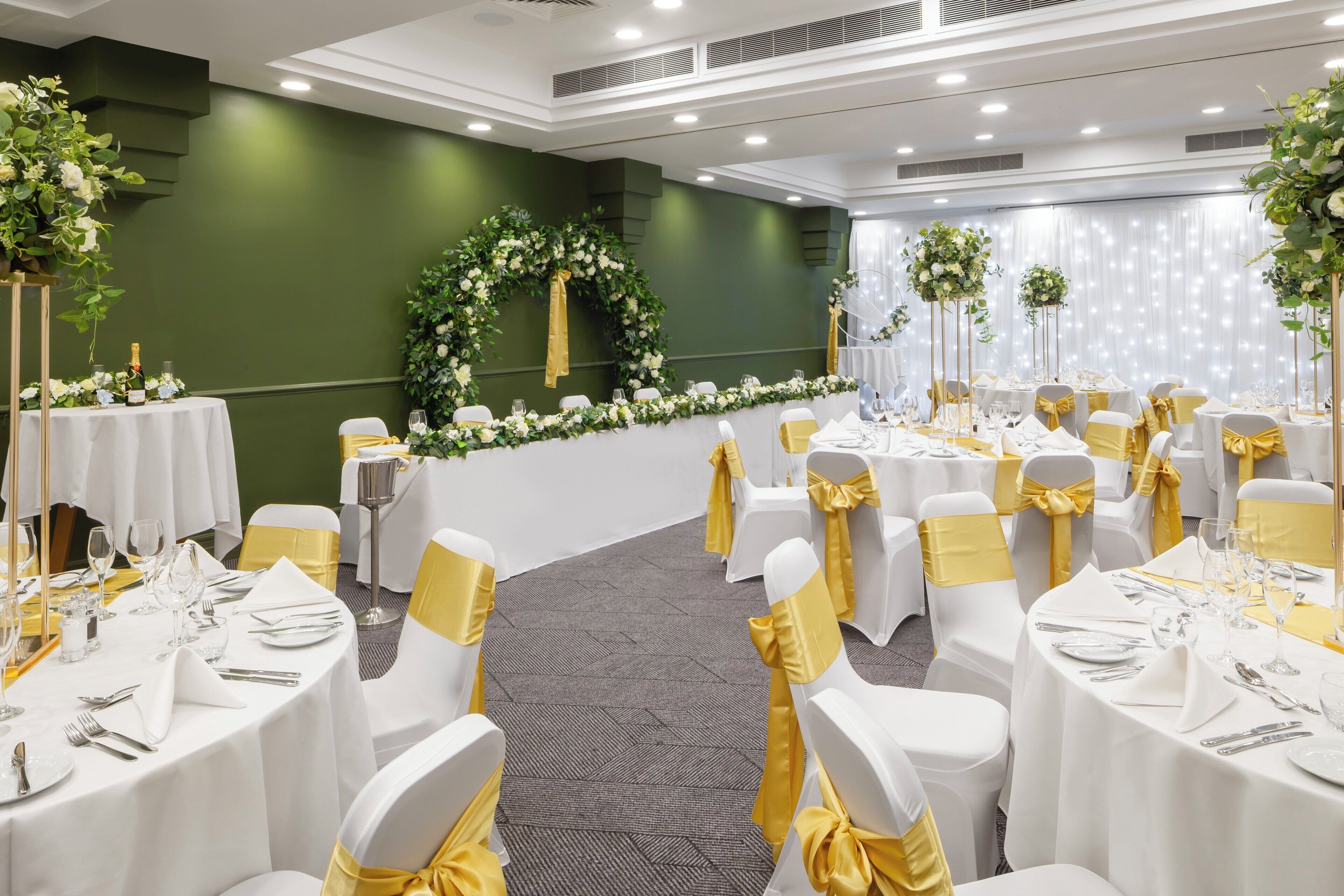Un salón de banquetes con mesas redondas, sillas blancas con cintas doradas y decoración floral.