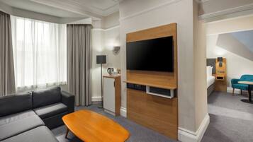 Una habitación de hotel moderna con un sofá gris, una mesa de centro de madera, televisor de pantalla plana y una pequeña zona de kitchenette.