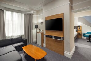 A modern hotel room with a grey sofa, wooden coffee table, flat-screen TV, and a small kitchenette area.