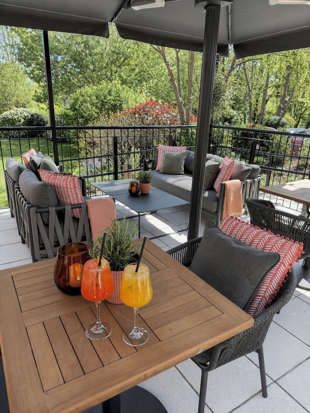 Un patio al aire libre con una mesa de madera, dos bebidas y una zona de estar con cojines y una planta en maceta.