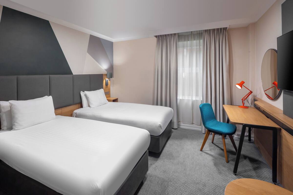 Ein Hotelzimmer mit zwei Betten, einem Schreibtisch, einem Stuhl, einem Fernseher und einem Fenster mit Vorhängen.