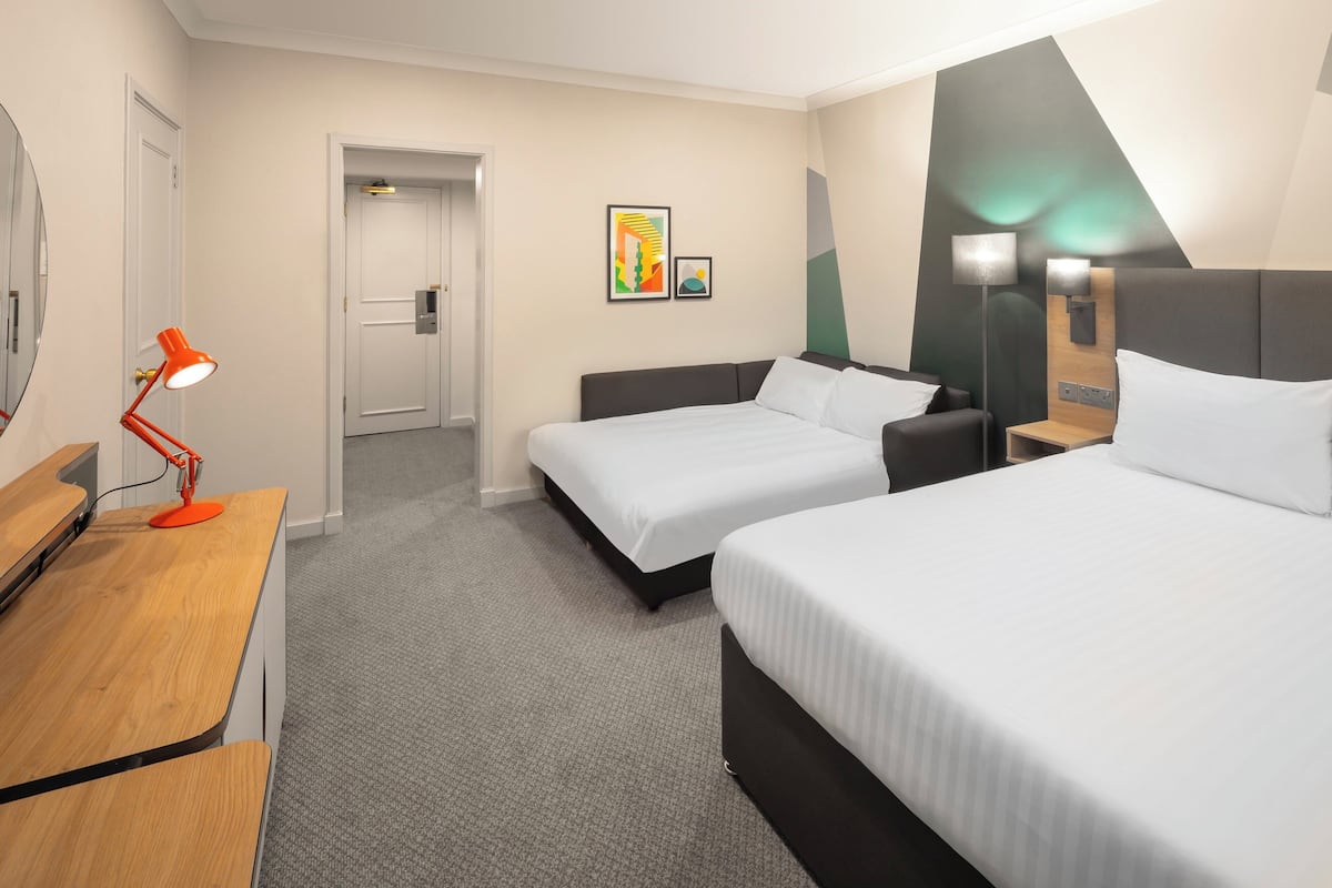Ein Hotelzimmer mit zwei Betten, einem Schreibtisch mit Lampe und einer Tür zu einem weiteren Raum.