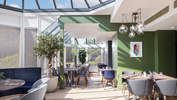 Un espace repas moderne doté d’un haut plafond, de grandes fenêtres et d’un puits de lumière. On y trouve plusieurs tables et chaises disposées pour accueillir les convives.