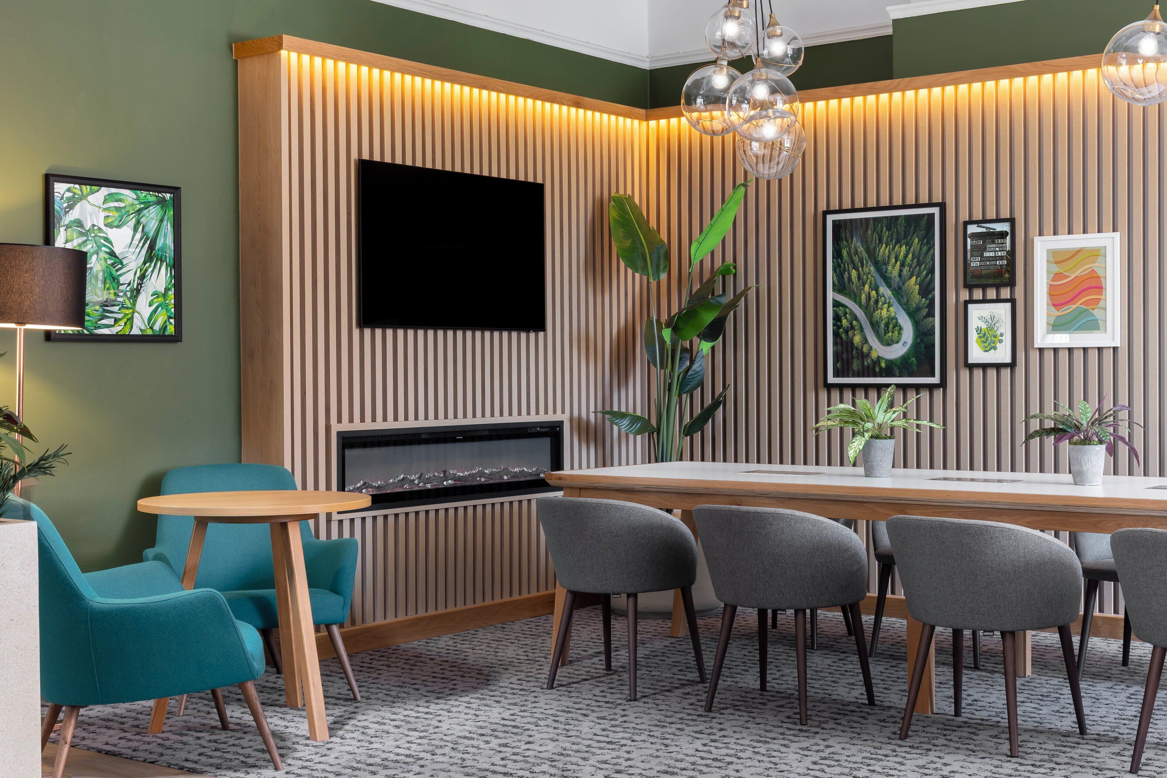 A modern dining area with a wooden table, grey upholstered chairs, and a flat-screen TV mounted on a slatted wall.