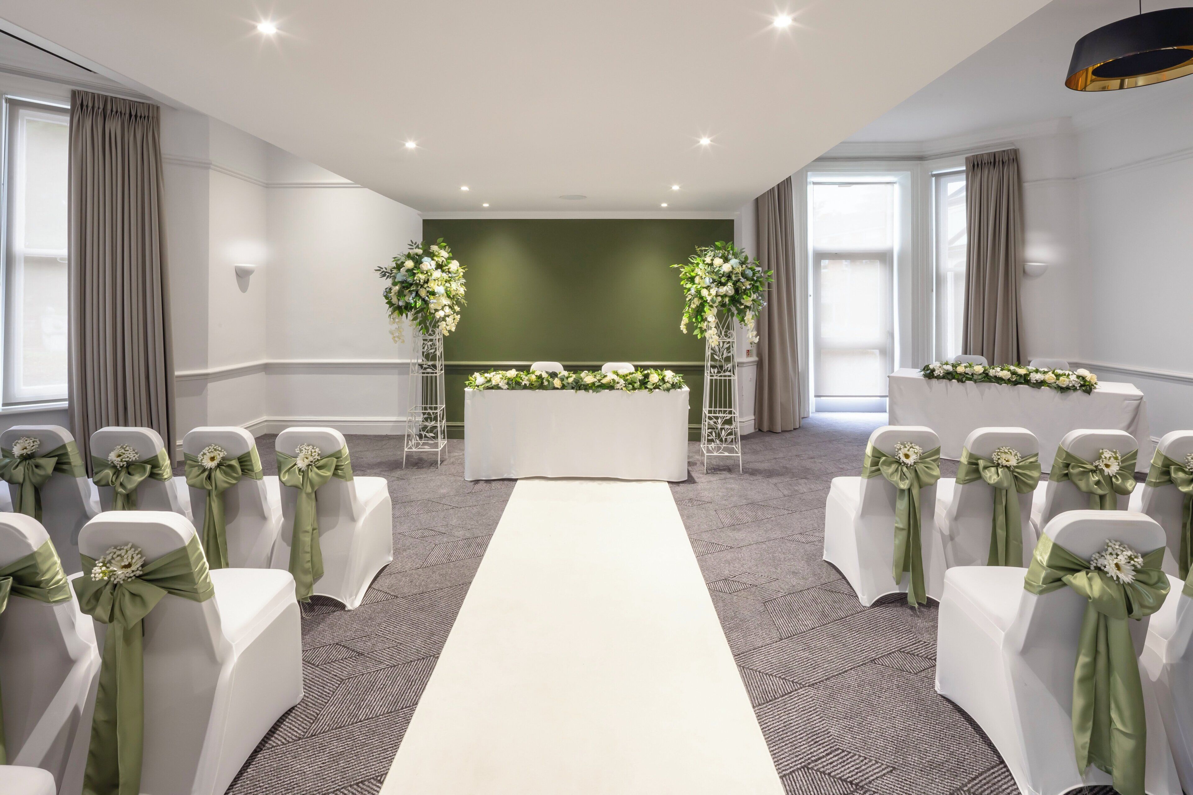 A room with a white aisle, white chairs with green sashes, a white table with floral arrangements, and a green accent wall.
