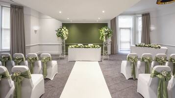 A room with a white aisle, white chairs with green sashes, a white table with floral arrangements, and a green accent wall.