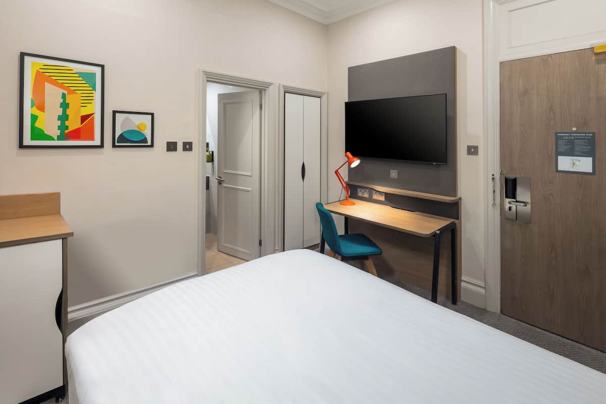 Ein Hotelzimmer mit Bett, Schreibtisch mit Stuhl, Fernseher und Kunstwerk an der Wand.