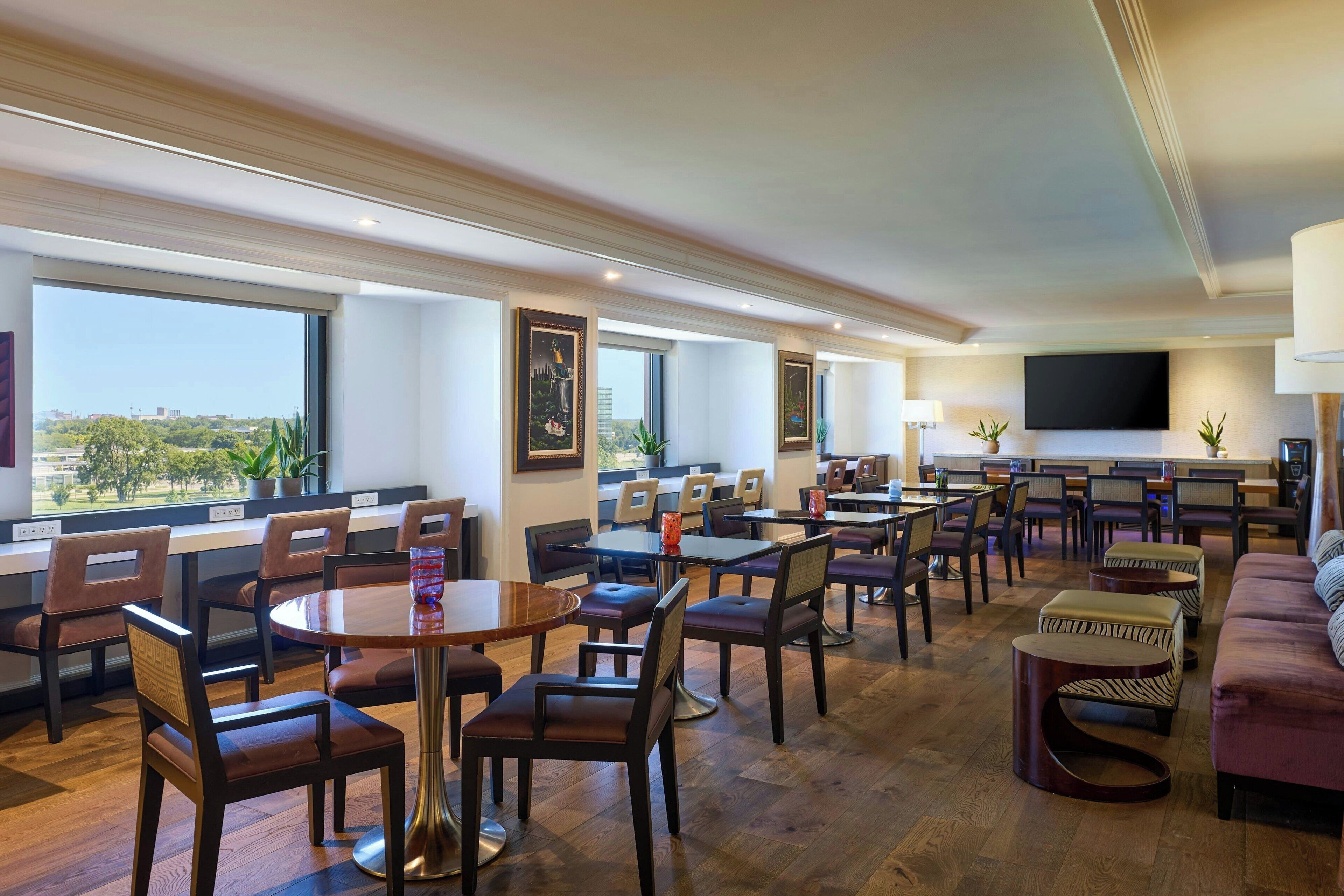 Un hall d’hôtel spacieux, doté de plusieurs espaces de détente, notamment des tables et des chaises rondes, un canapé et un téléviseur fixé au mur.