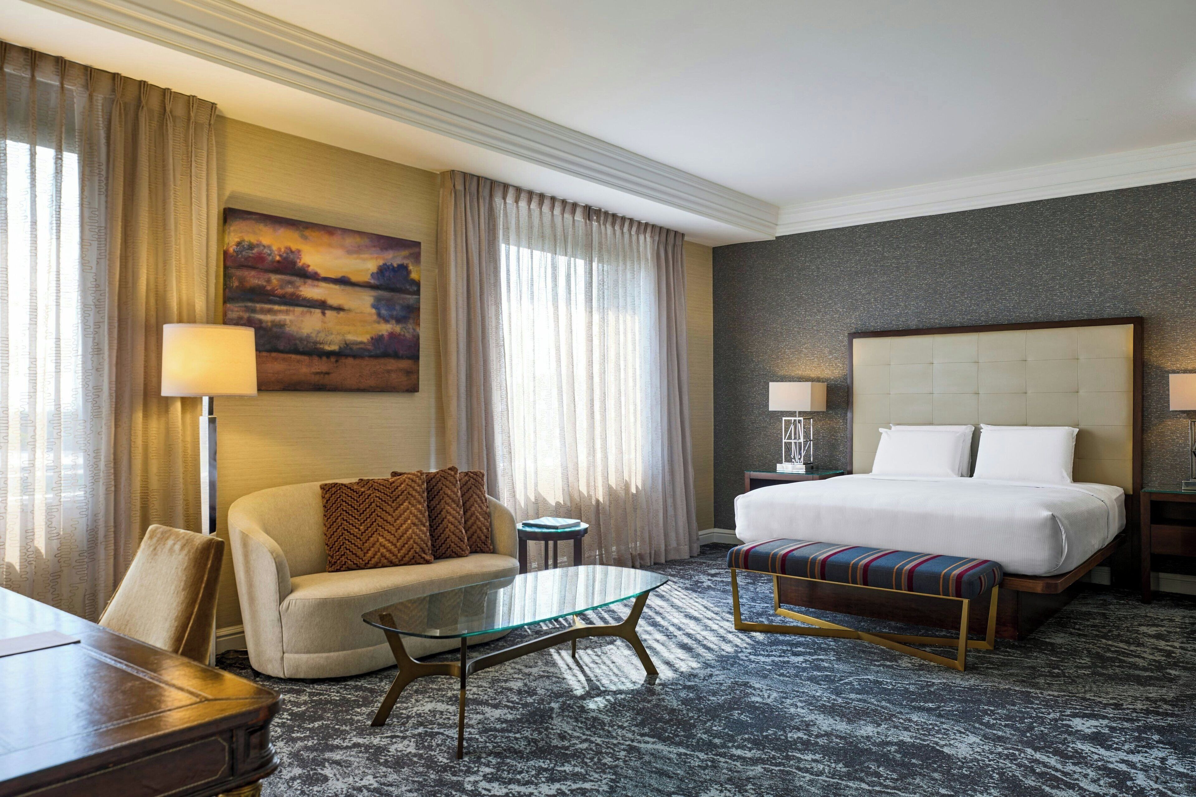 A hotel room with a bed, a sofa, a glass-top coffee table, and a desk.
