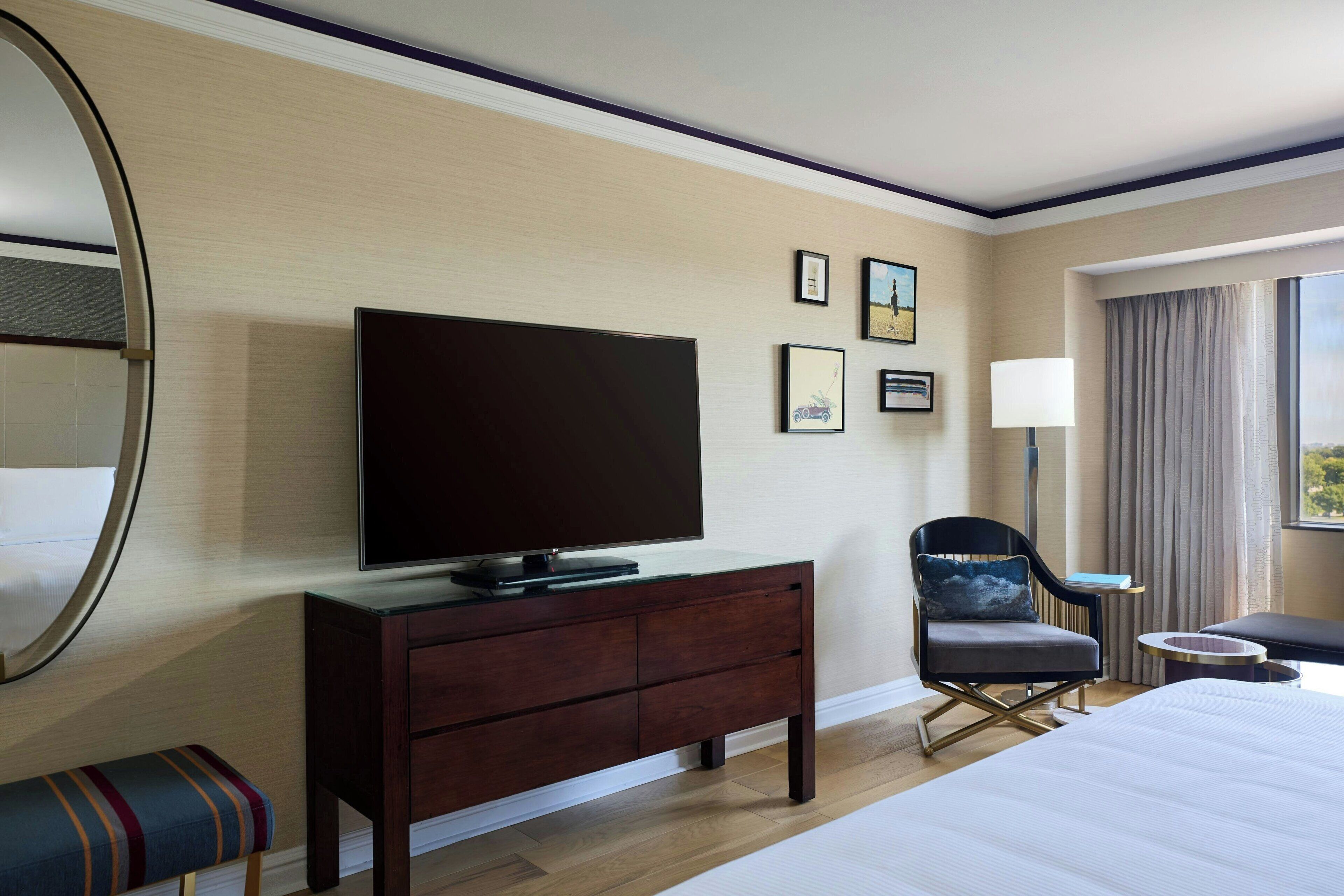 A hotel room with a large bed, a TV on a wooden dresser, a chair, a small table, and a window with curtains.