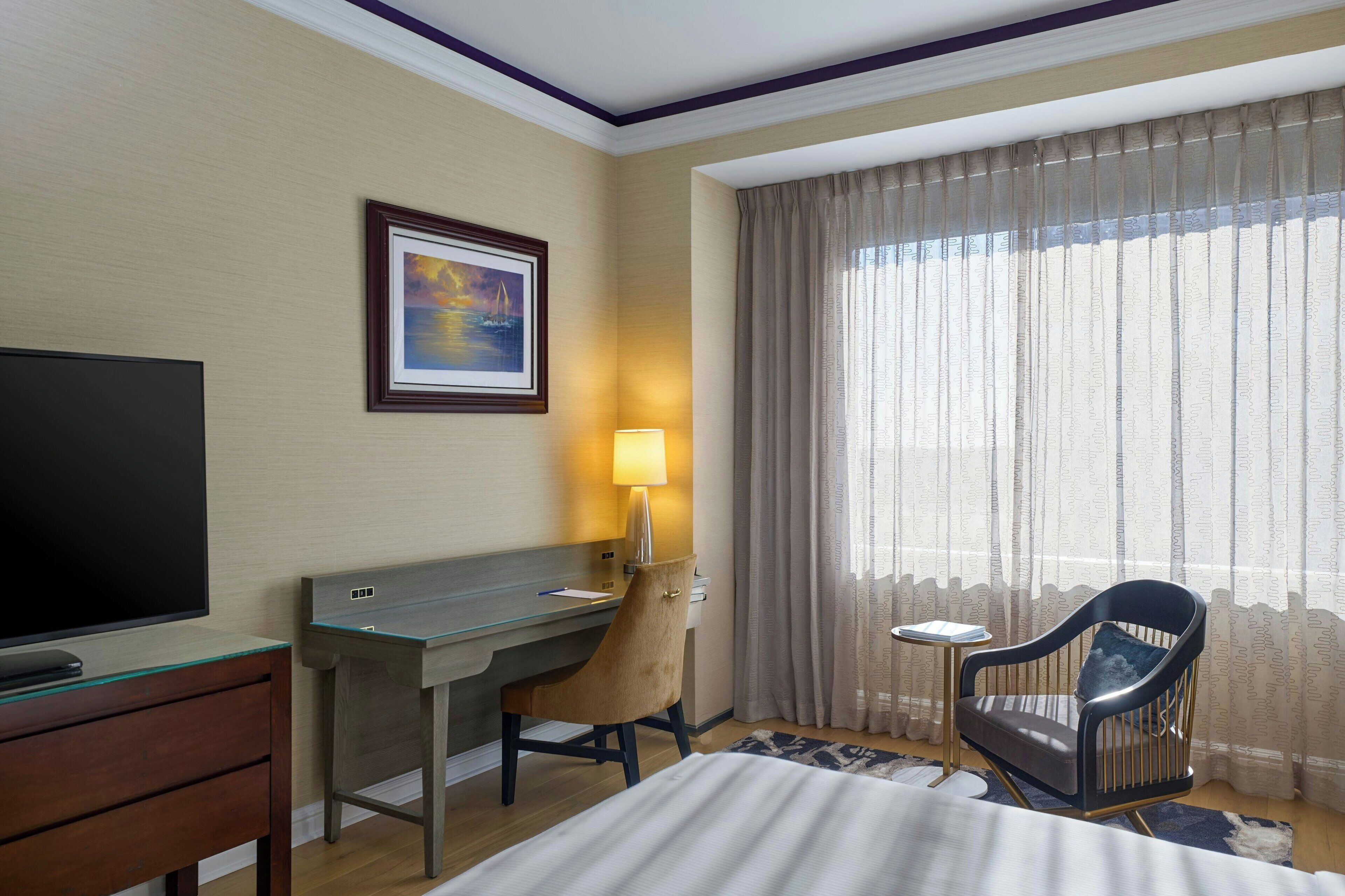 Una habitación de hotel con una cama, un escritorio con silla, televisión, lámpara y ventana con cortinas.