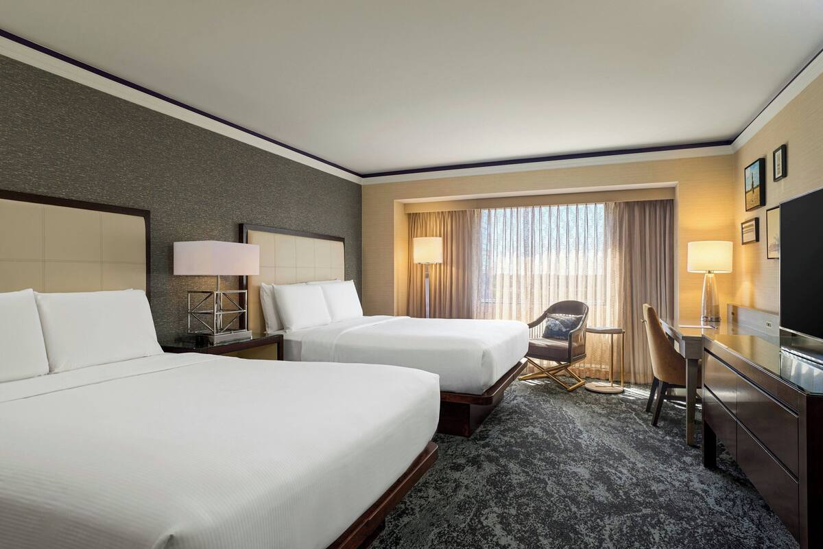 A hotel room with two beds, a desk, a chair, a TV, and a window with curtains.