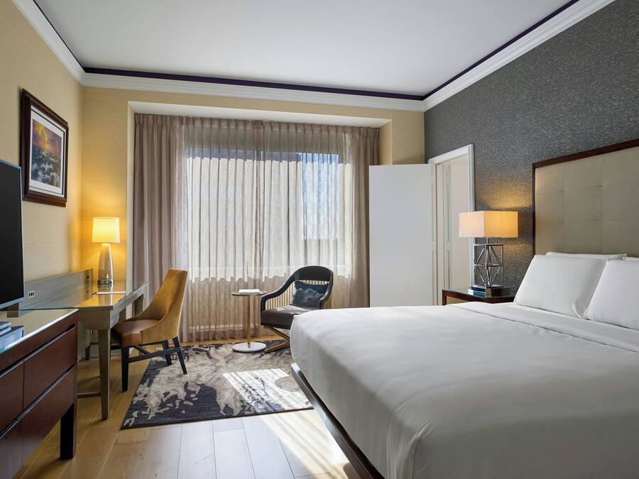 Ein Hotelzimmer mit einem großen Bett, einem Schreibtisch mit Stuhl, einem Fernseher und einem Fenster mit Vorhängen.