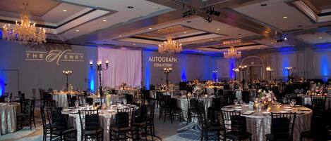A banquet hall with round tables, black chairs, chandeliers, and a sign reading "The Henry Autograph Collection".