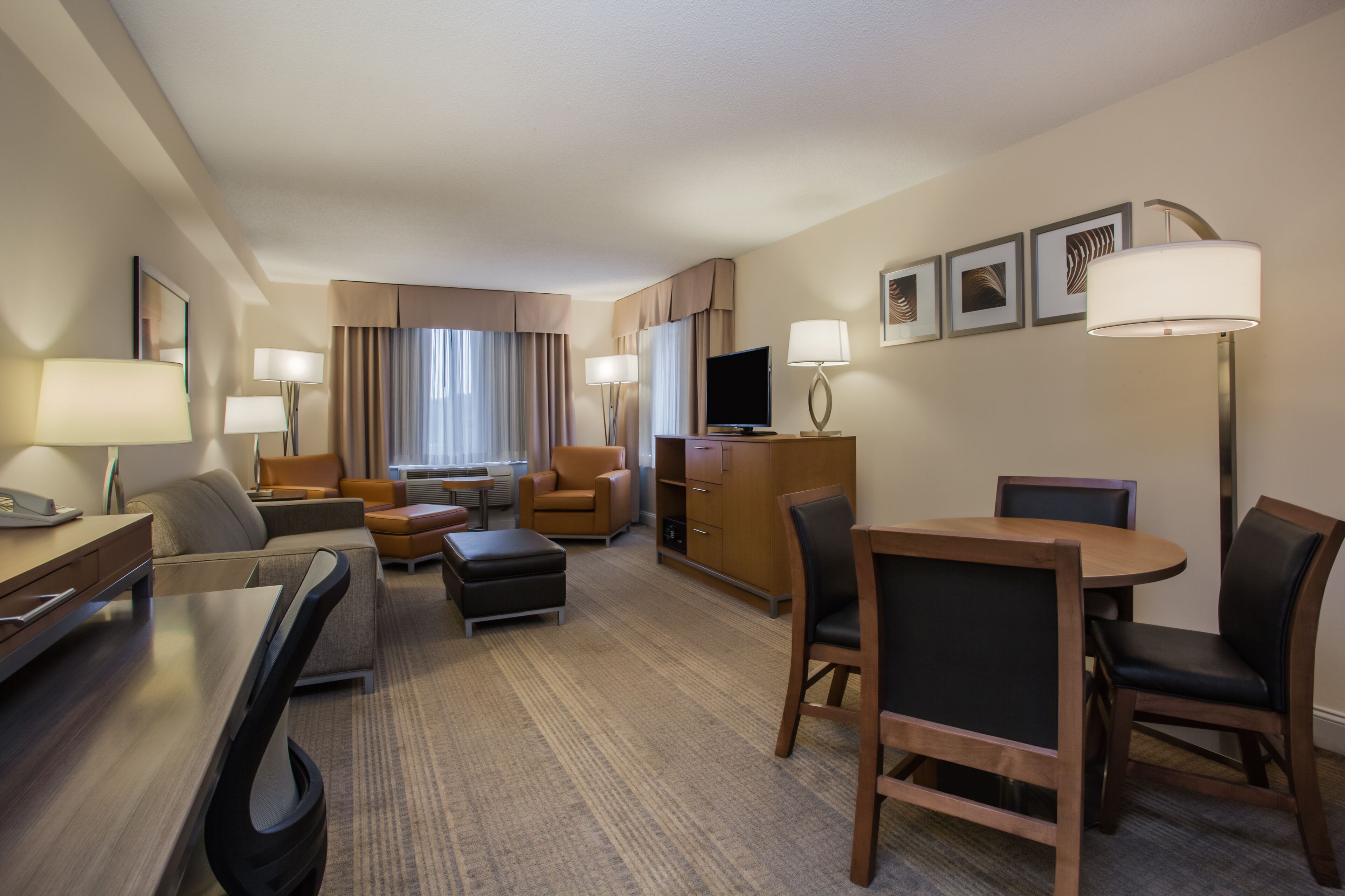 Habitación de hotel con sofá, sillón, reposapiés, mesa de comedor, sillas, televisión y escritorio con lámpara.