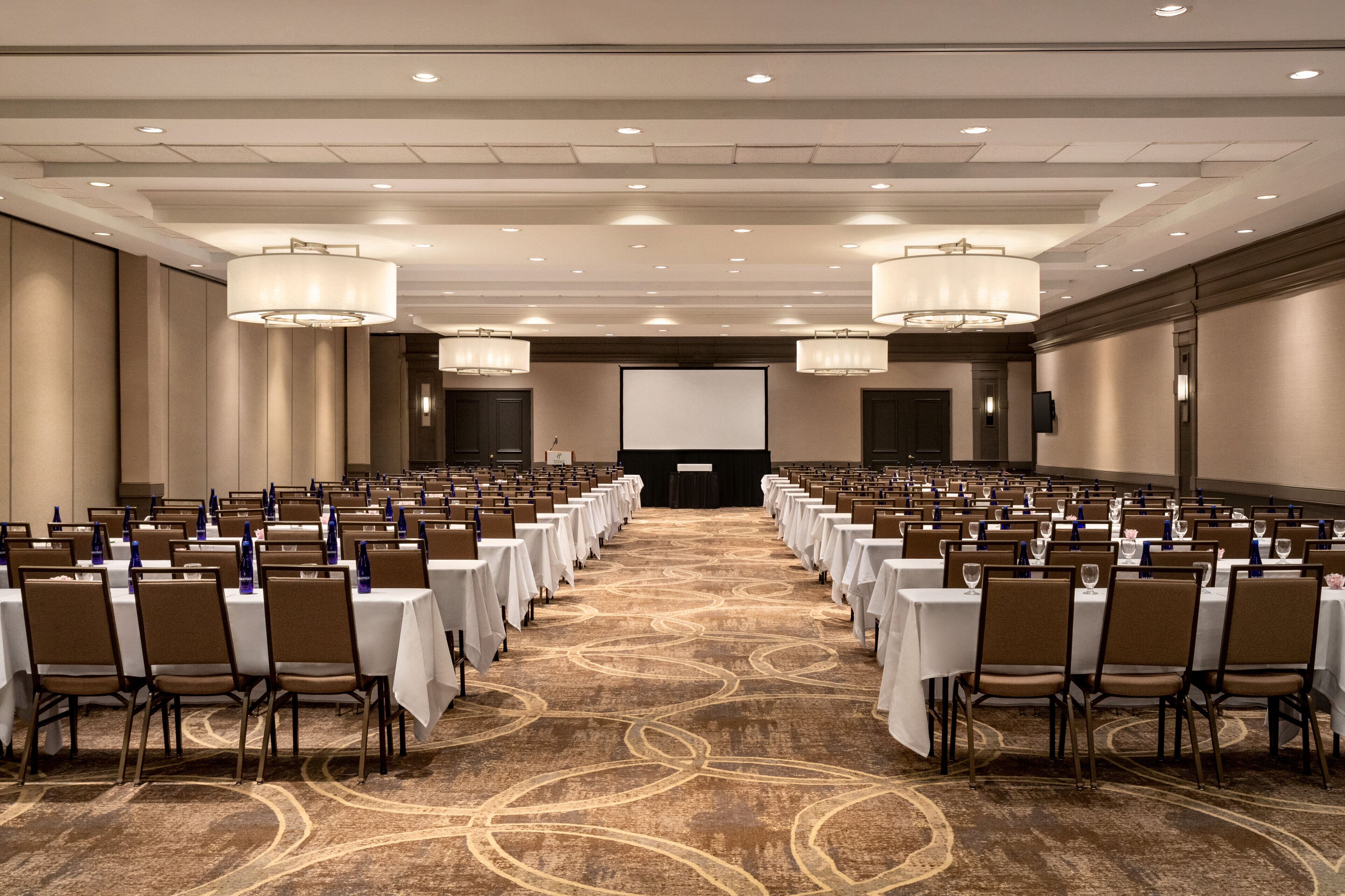Un salón de conferencias con filas de mesas y sillas, una pantalla grande al frente y candelabros.