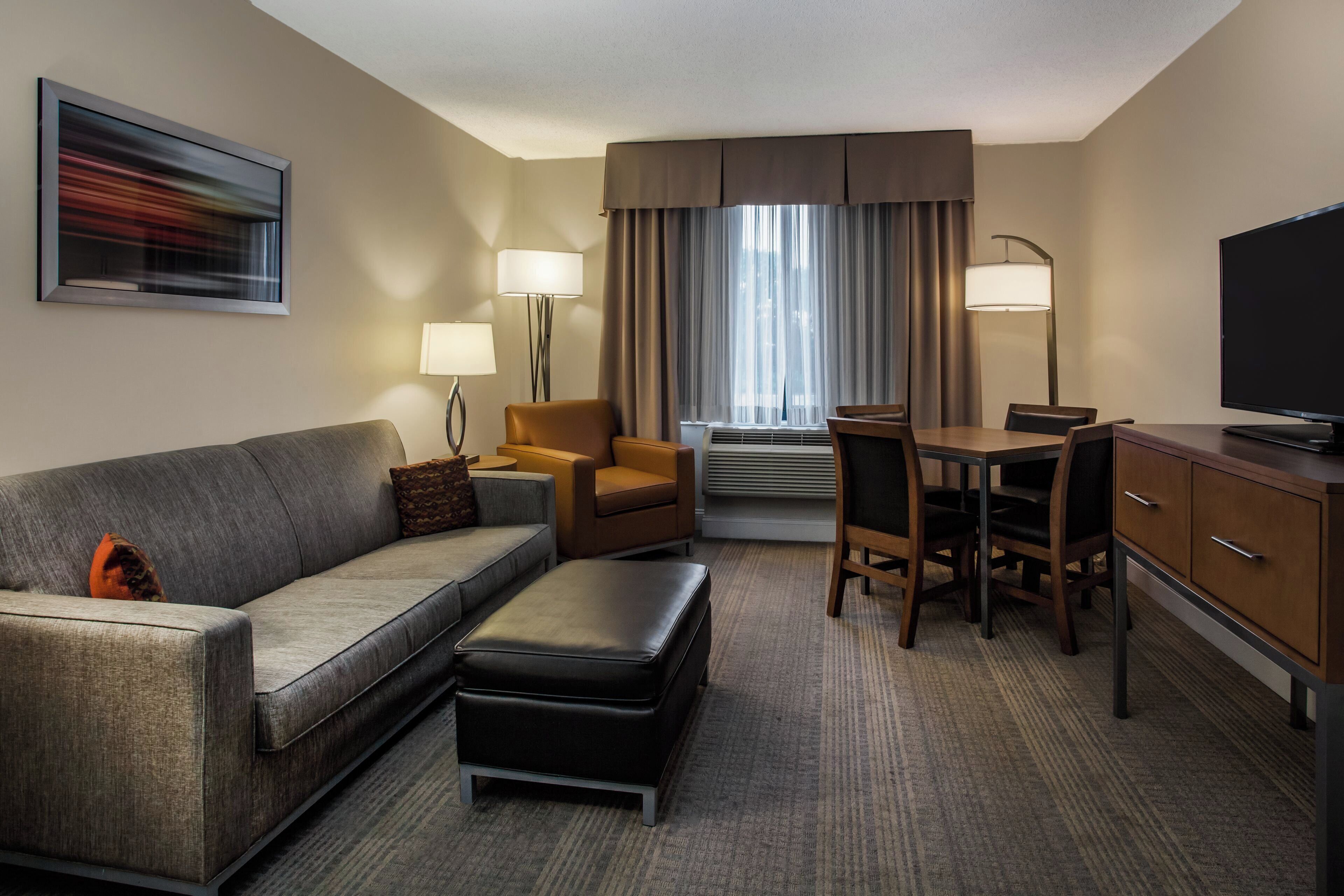 A hotel room with a sofa, armchair, ottoman, desk, chairs, lamps, and a television.