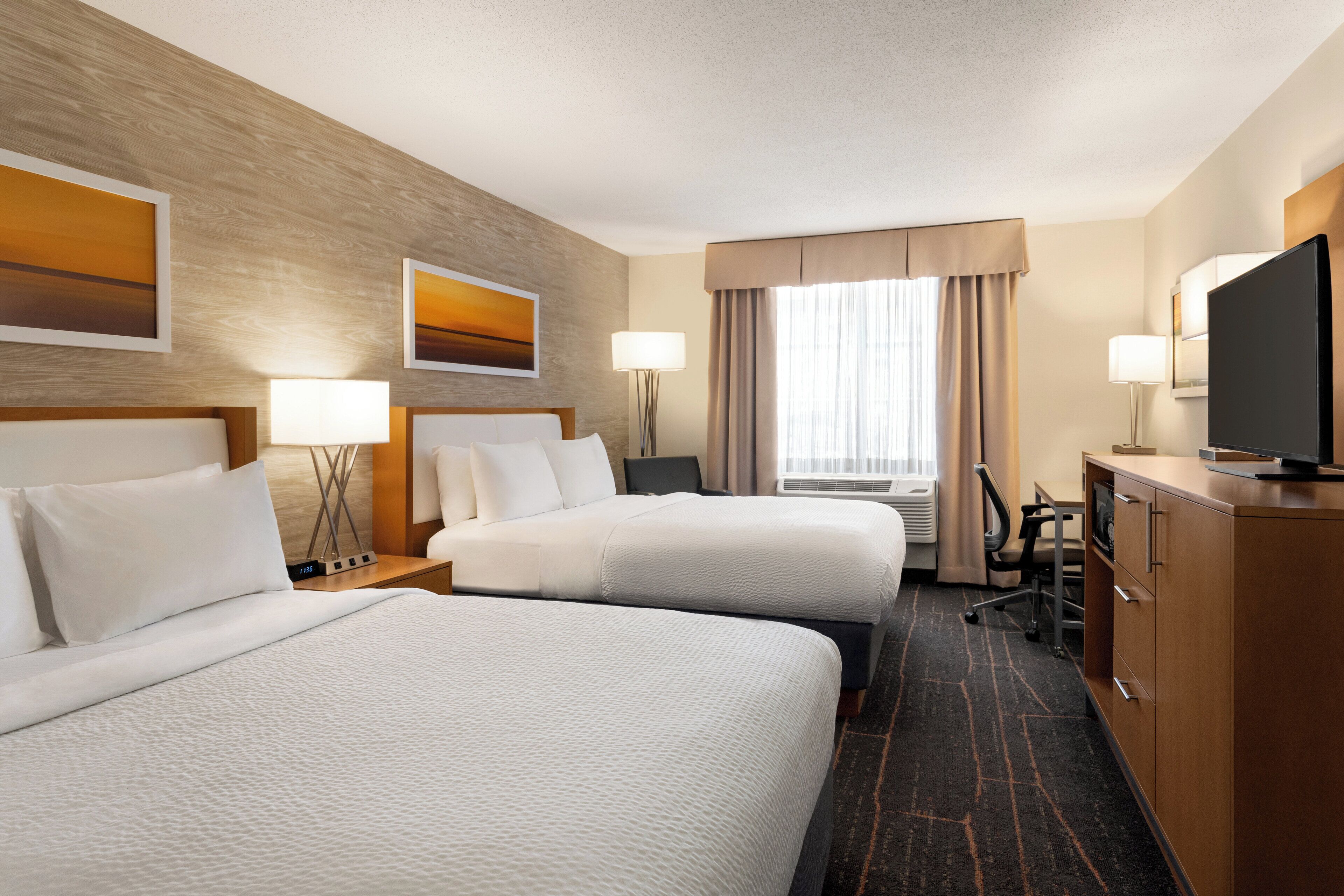 A hotel room with two beds, a desk, a television, and a large window with curtains.