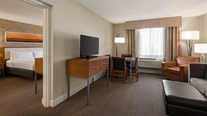 Habitación de hotel con una cama, un escritorio con televisor, una silla, un sofá y una ventana con cortinas.